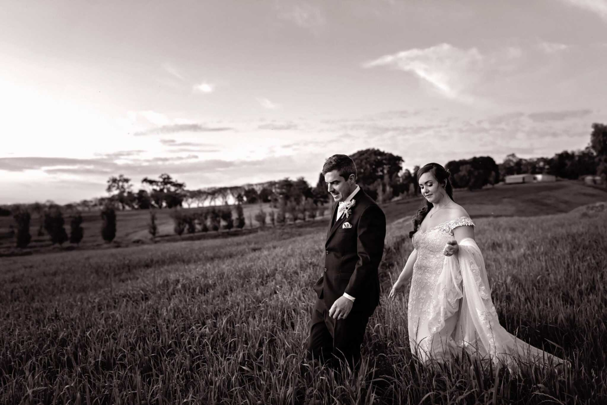 Bride and groom walk through a grassy field at sunset in a romantic outdoor wedding portrait.