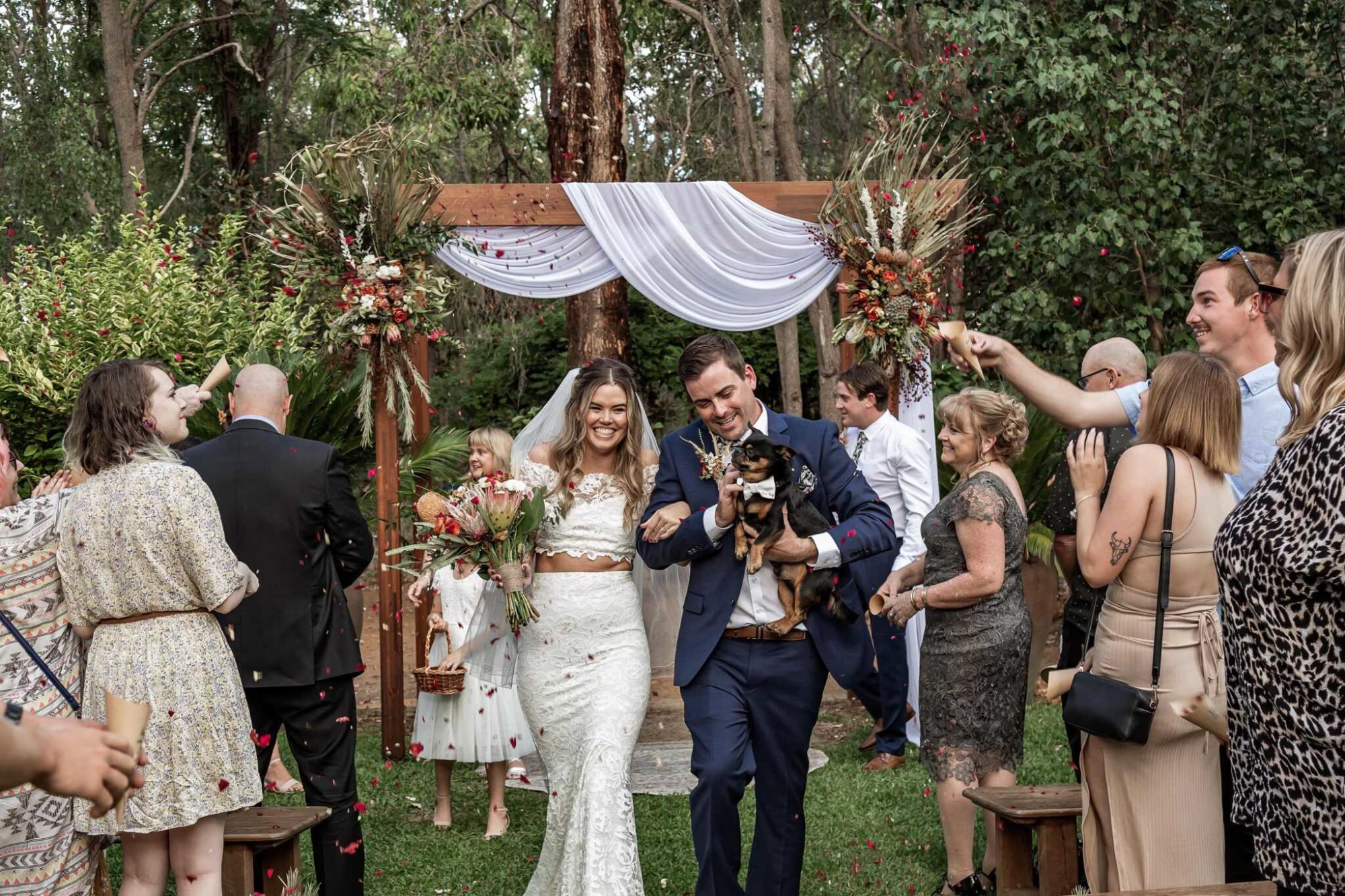 Newlywed couple walks down an outdoor aisle with guests tossing petals under a rustic floral arbor.