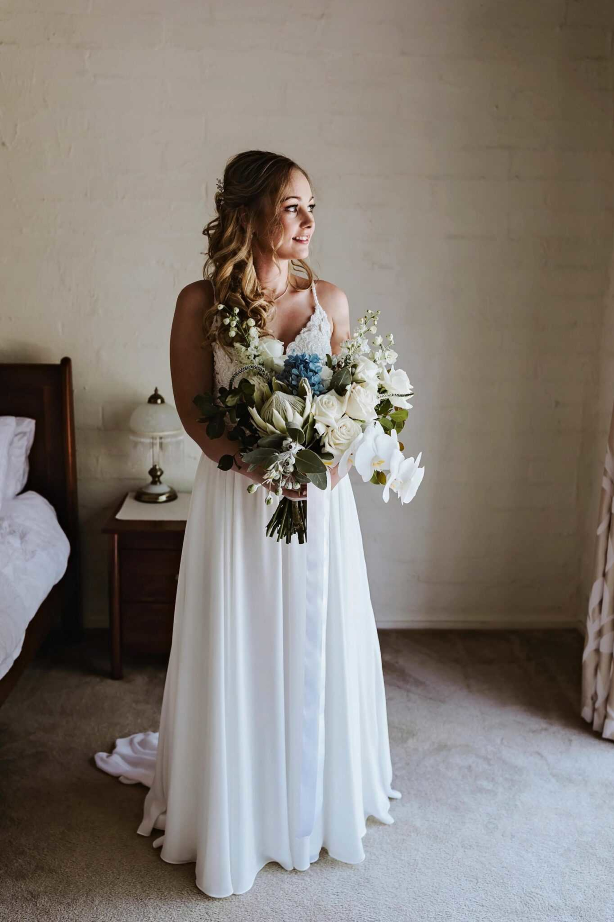 Bride in a white gown holding a lush bouquet, standing in a softly lit bedroom and looking to the side.