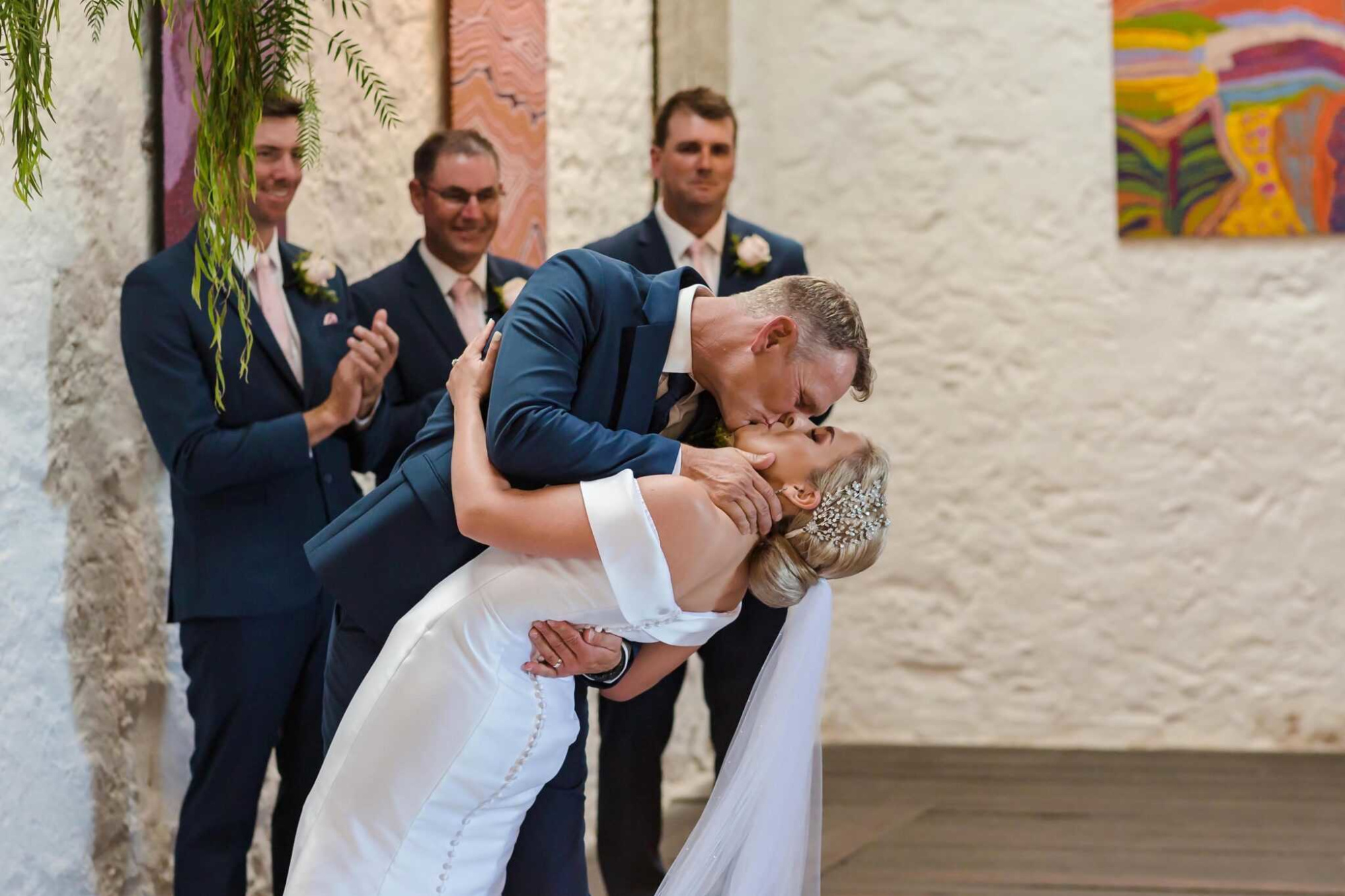 Groom dips and kisses bride during indoor wedding ceremony as groomsmen smile in the background.