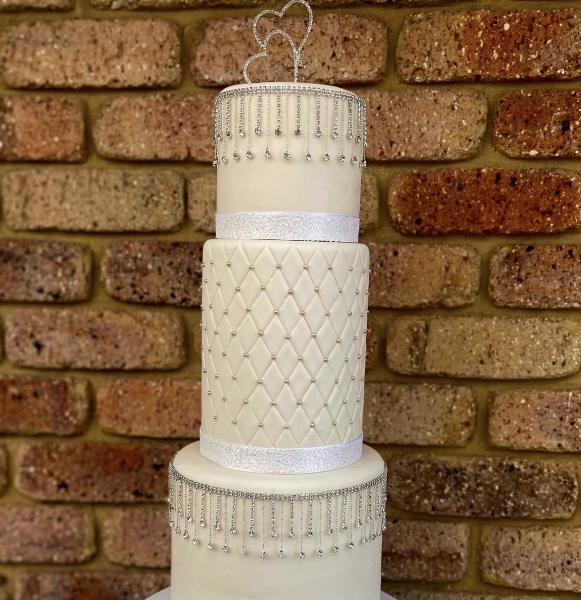 Tall three-tier white wedding cake with crystal fringe and double heart topper against a brick wall backdrop.