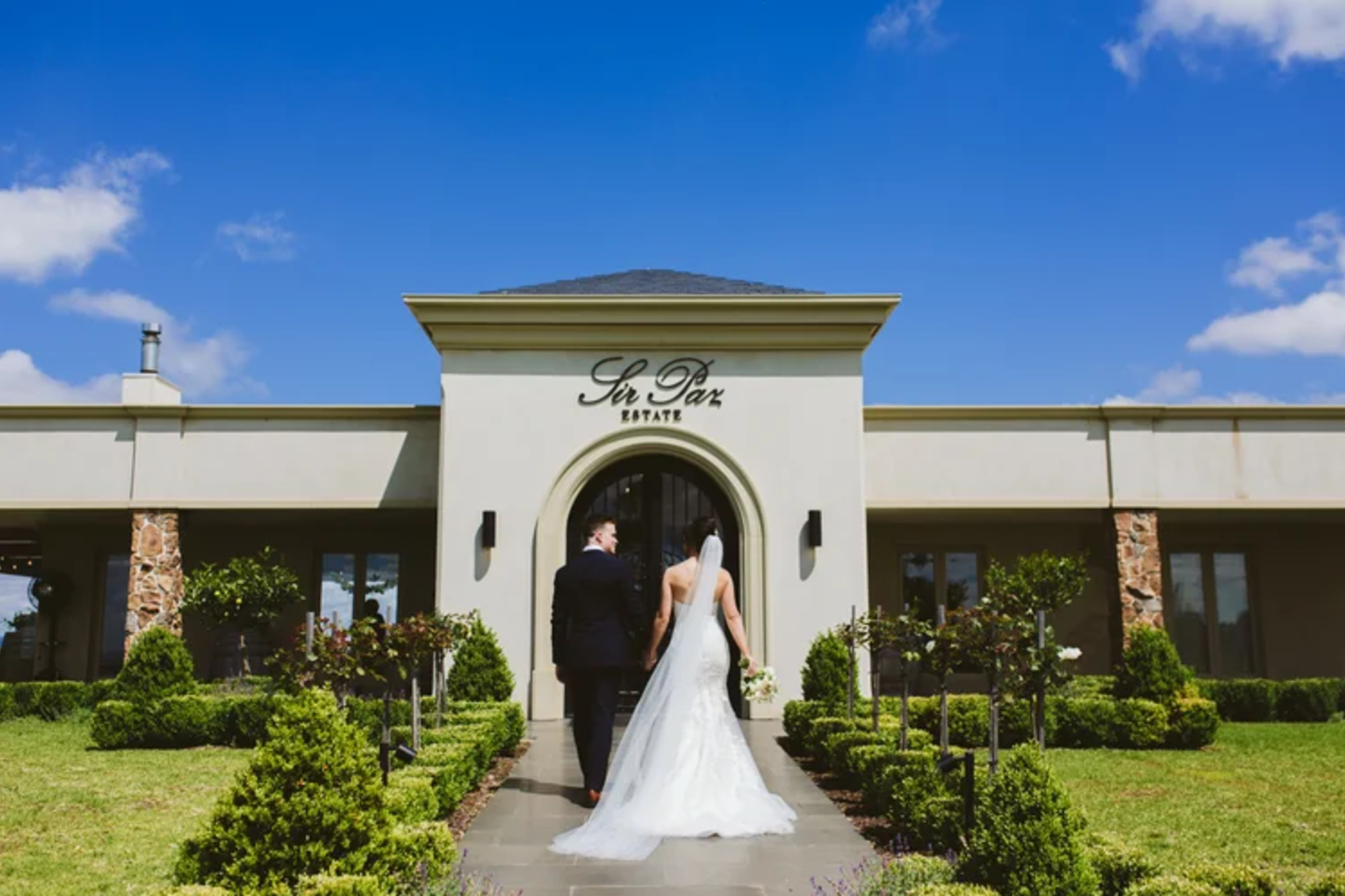 Bride and groom walk hand in hand toward an elegant estate entrance on a sunny day.