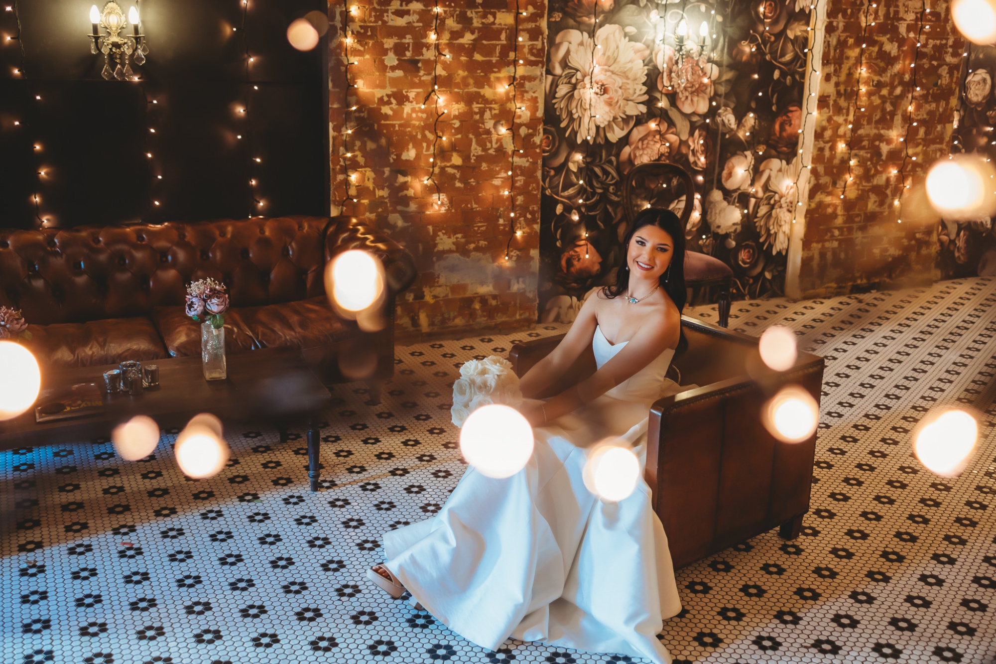 Bride in a white gown poses in a cozy lounge with fairy lights and floral wallpaper.