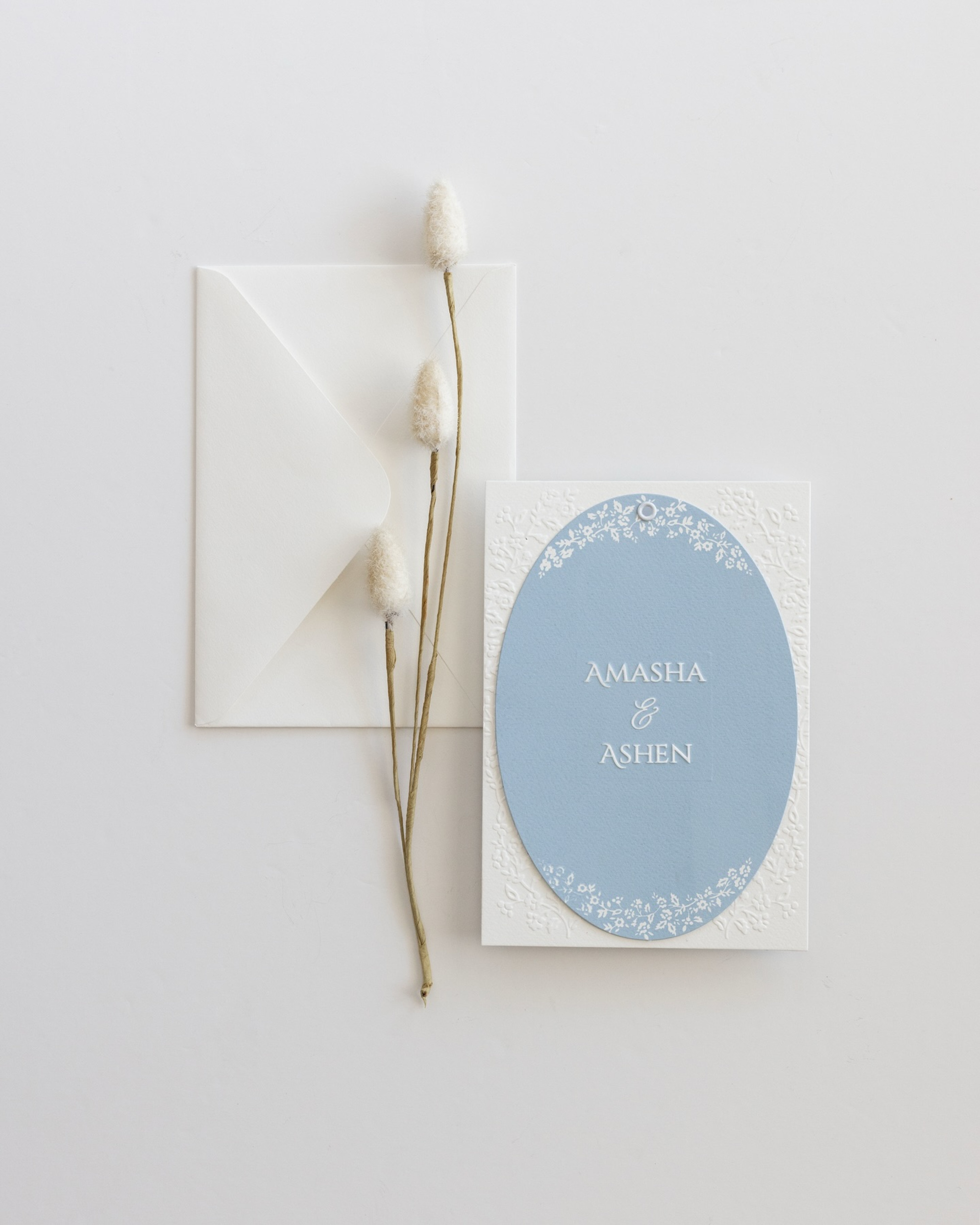 Minimalist blue and white wedding invitation suite with dried flowers and envelope on a white background.