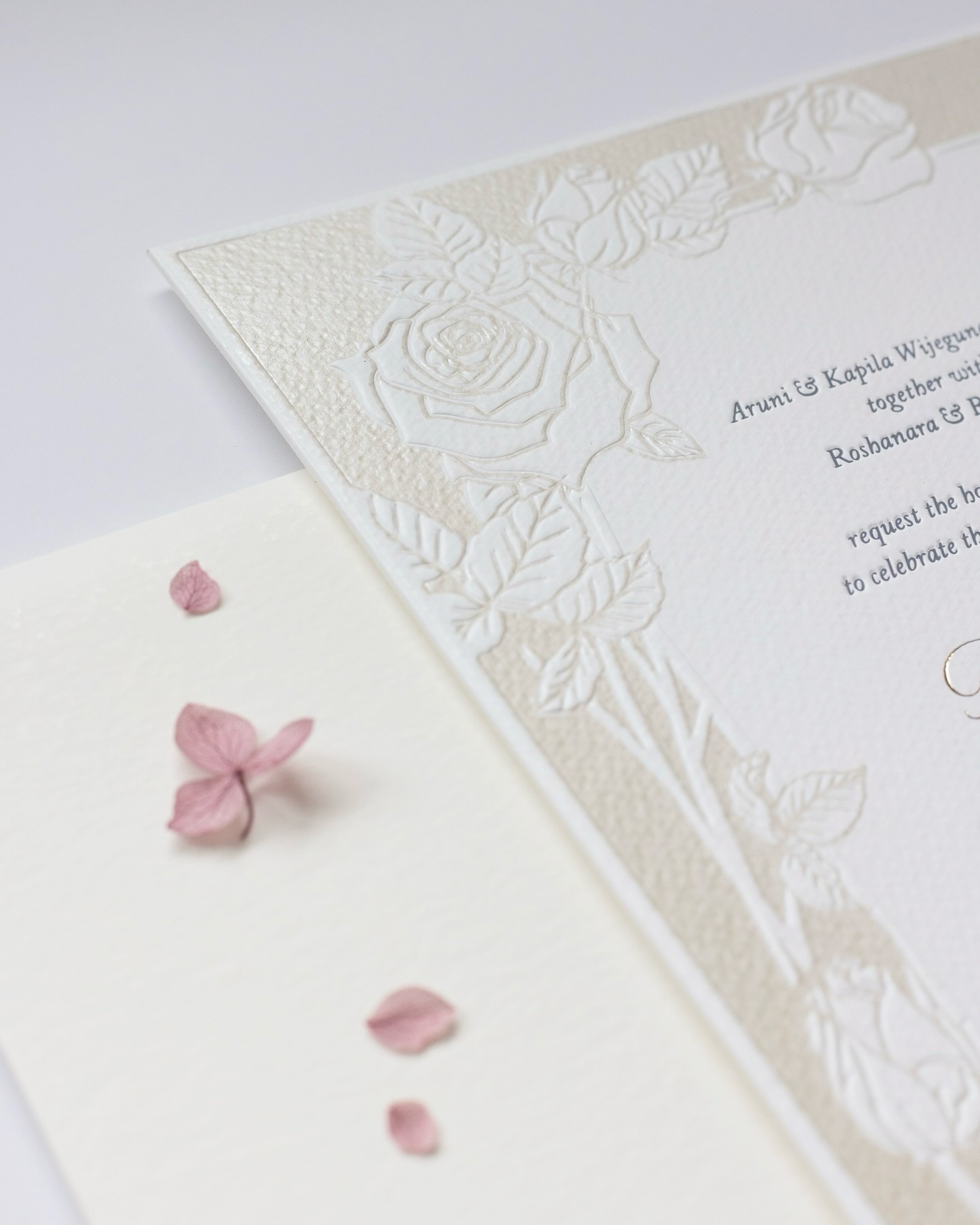 Elegant embossed floral wedding invitation with blush petals scattered beside it.