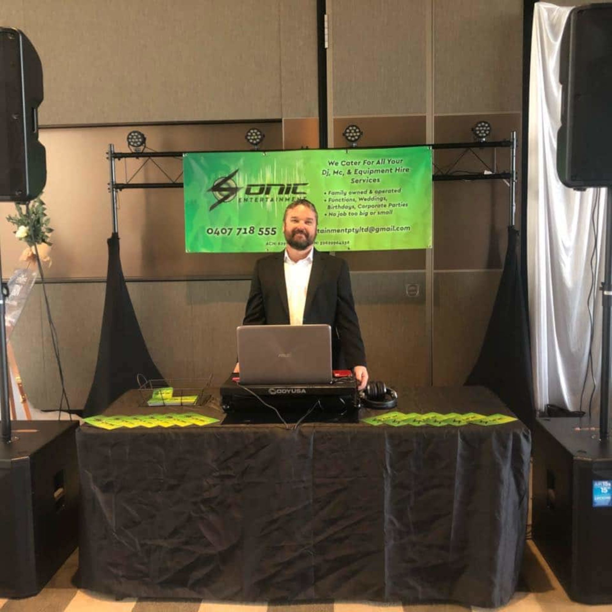 Wedding DJ in a suit standing behind a laptop and DJ booth with speakers and a green company banner.