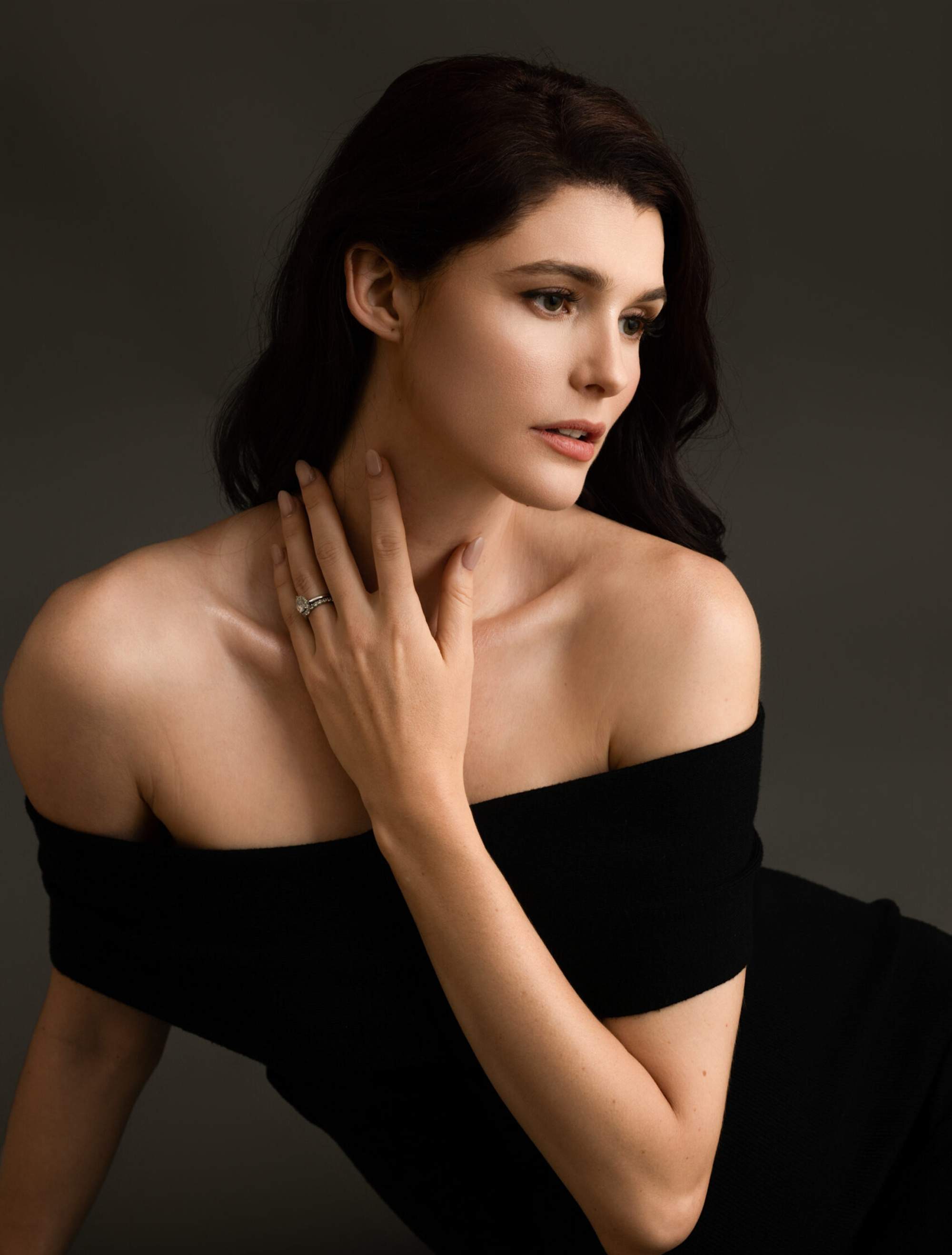 Elegant bridal portrait of a woman in a black off-shoulder dress showcasing her engagement ring against a dark background.