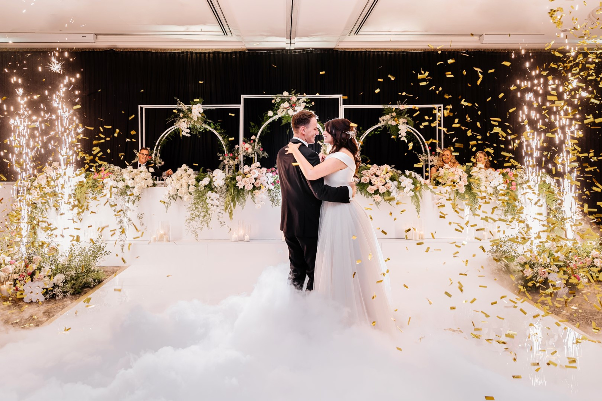 Bride and groom share their first dance amid sparklers, confetti, and lush floral decor on a cloud-like dance floor.