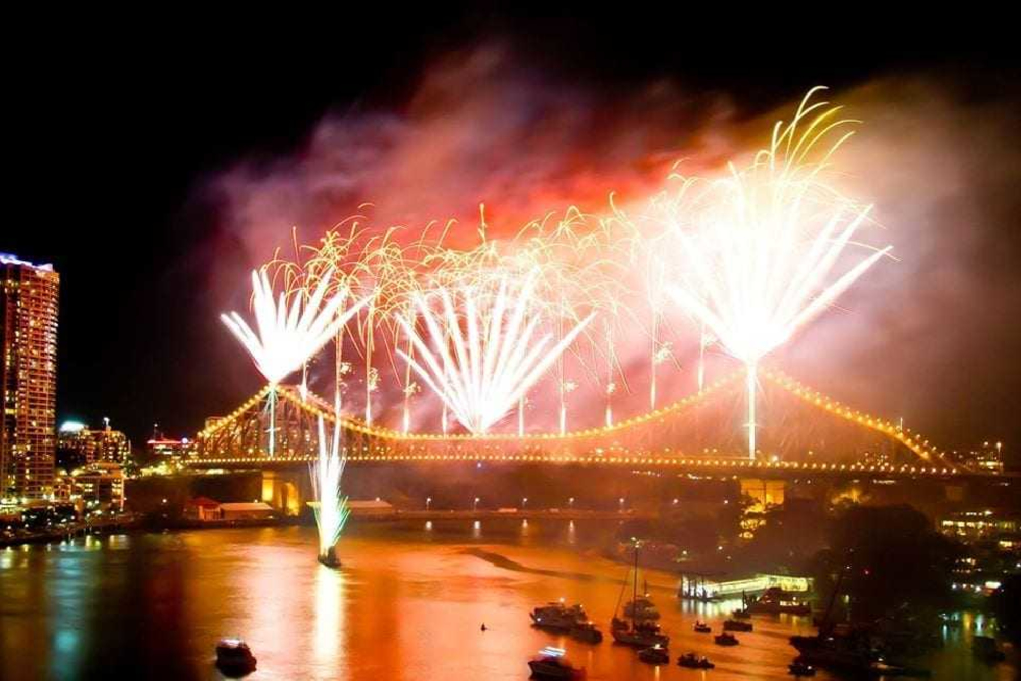 Bright fireworks explode over a lit city bridge and river at night during a wedding celebration.