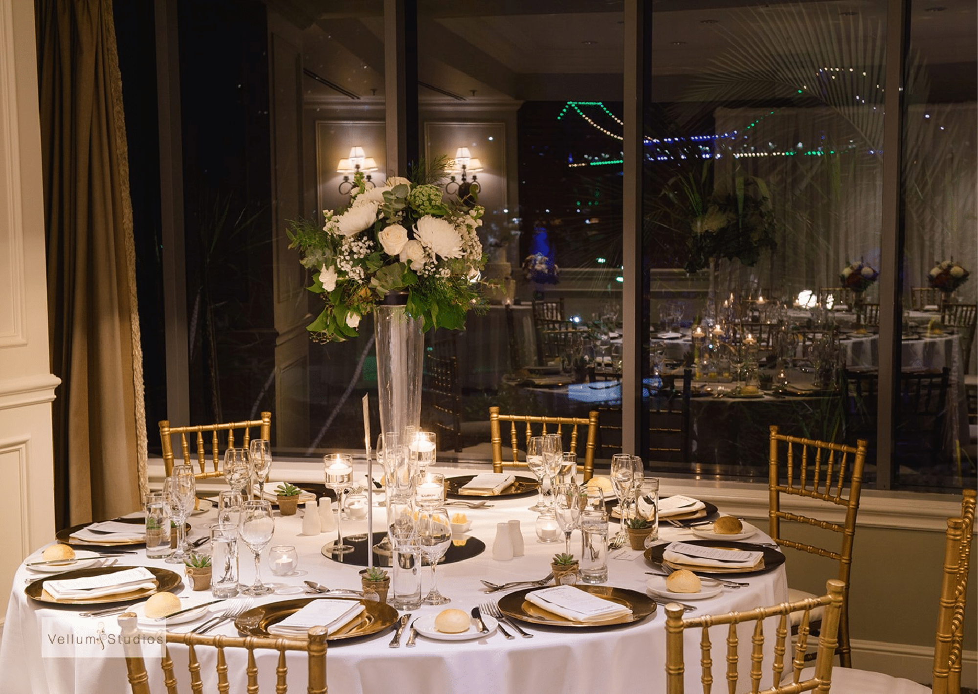 Elegant indoor wedding reception table with tall floral centerpiece, gold chairs, and candlelit place settings at night.