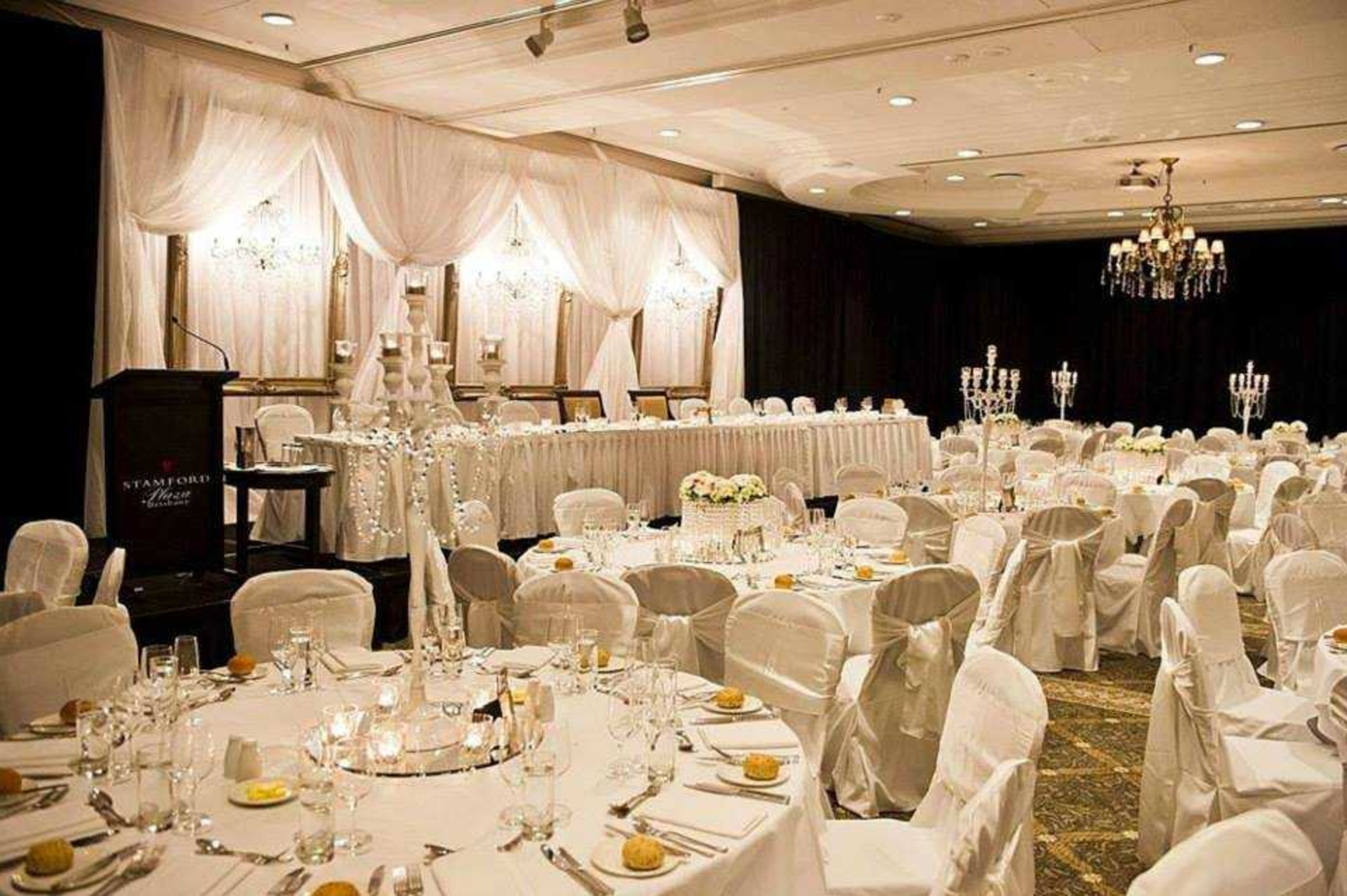 Elegant indoor wedding reception with draped backdrop, chandeliers, and round tables set with white linens and glassware.