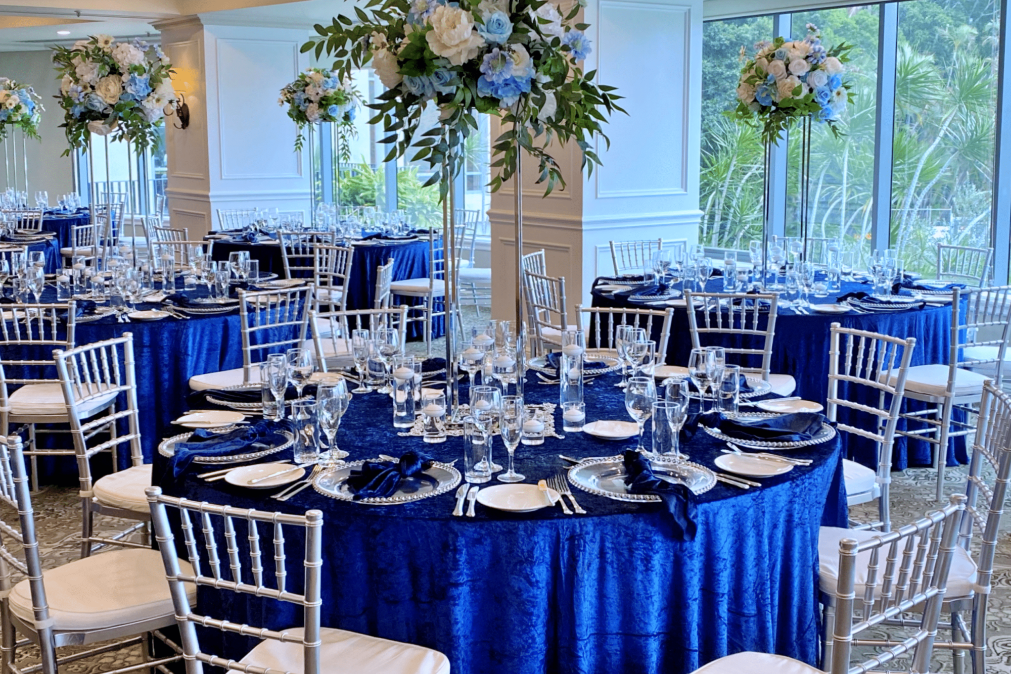 Elegant wedding reception ballroom with royal blue linens, tall floral centerpieces, and silver chairs set for guests.