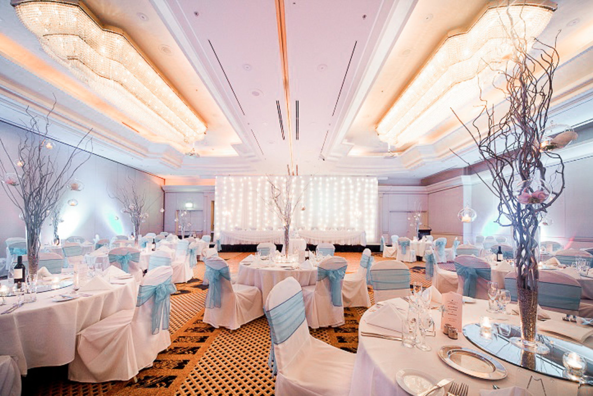 Elegant indoor wedding reception with round tables, blue chair sashes, and twinkling backdrop lighting.