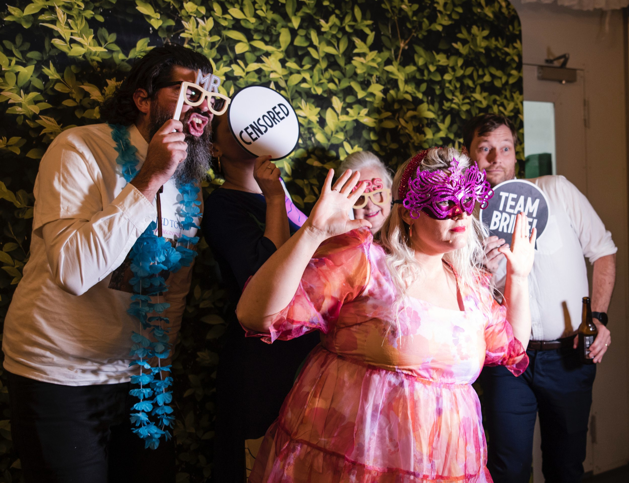 Wedding guests pose with playful photobooth props in front of a leafy backdrop.