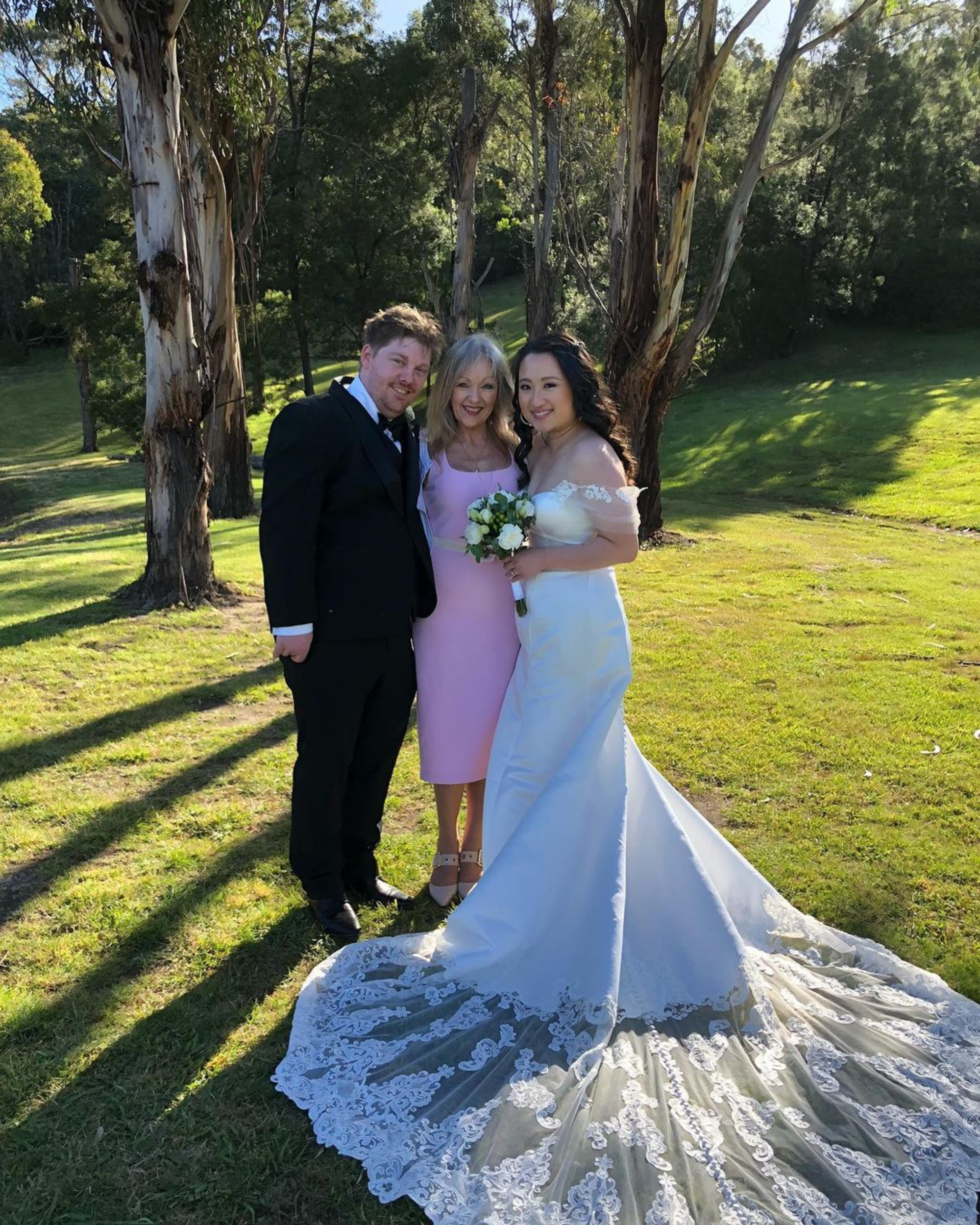 Bride, groom, and guest pose together outdoors in a sunlit grassy park with tall trees.
