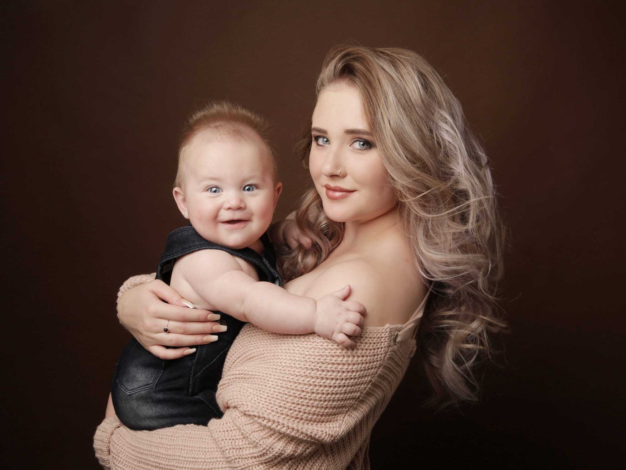 Studio portrait of a woman holding a smiling baby against a plain brown background.