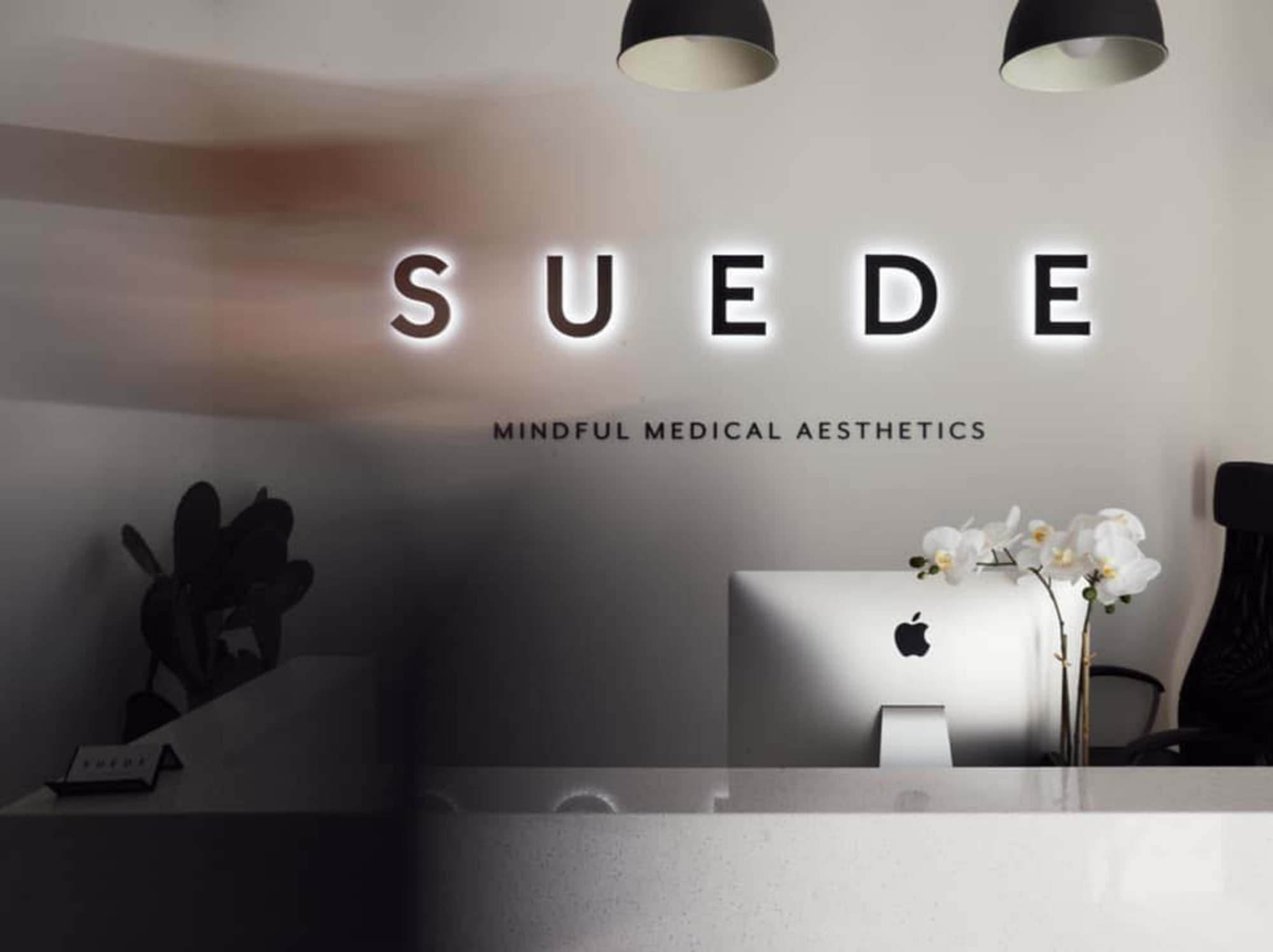 Modern aesthetic clinic reception with SUEDE backlit sign, sleek desk, and white orchids.