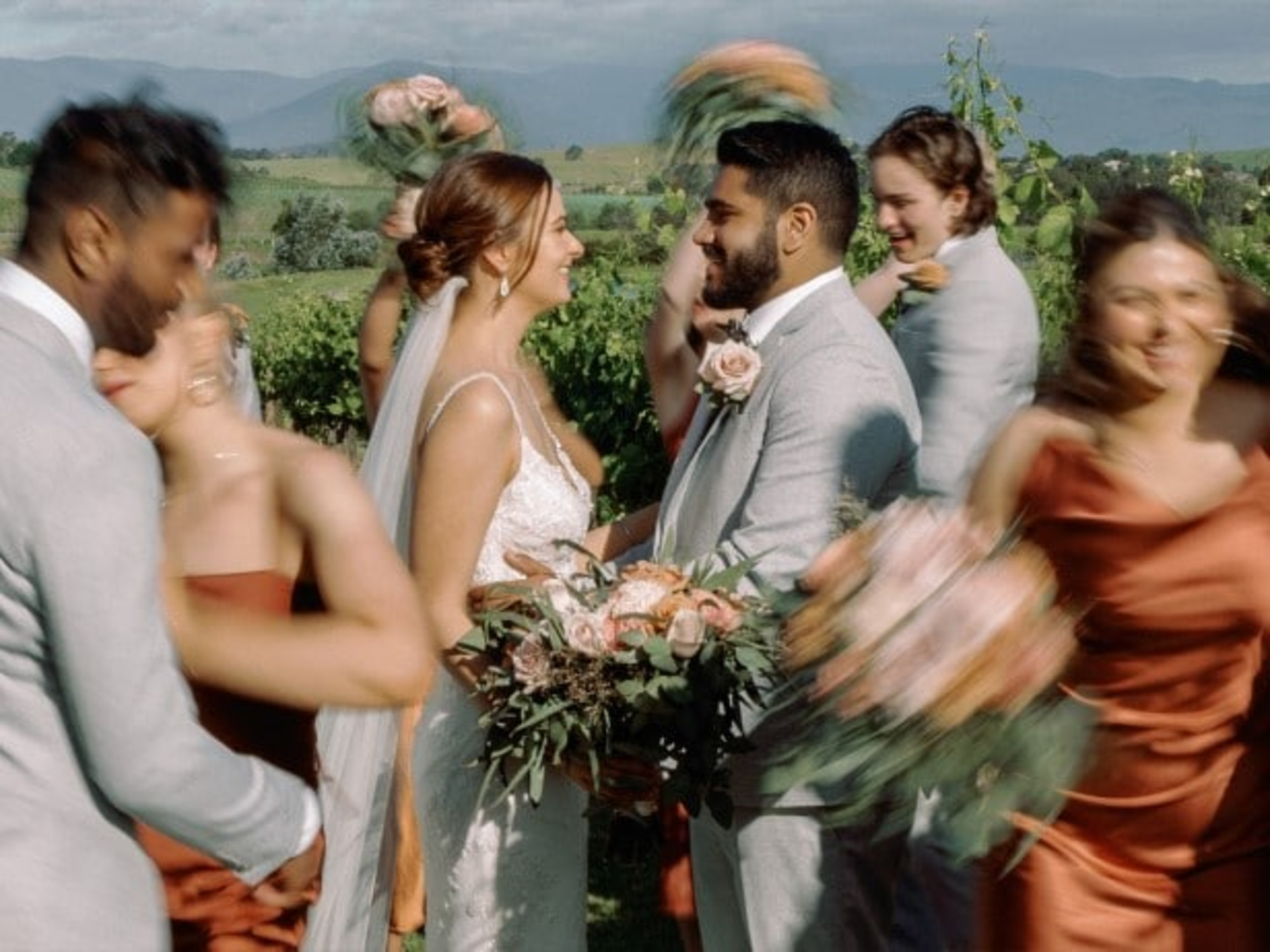 Bride and groom smile at each other surrounded by their wedding party in a vineyard with mountains in the background.