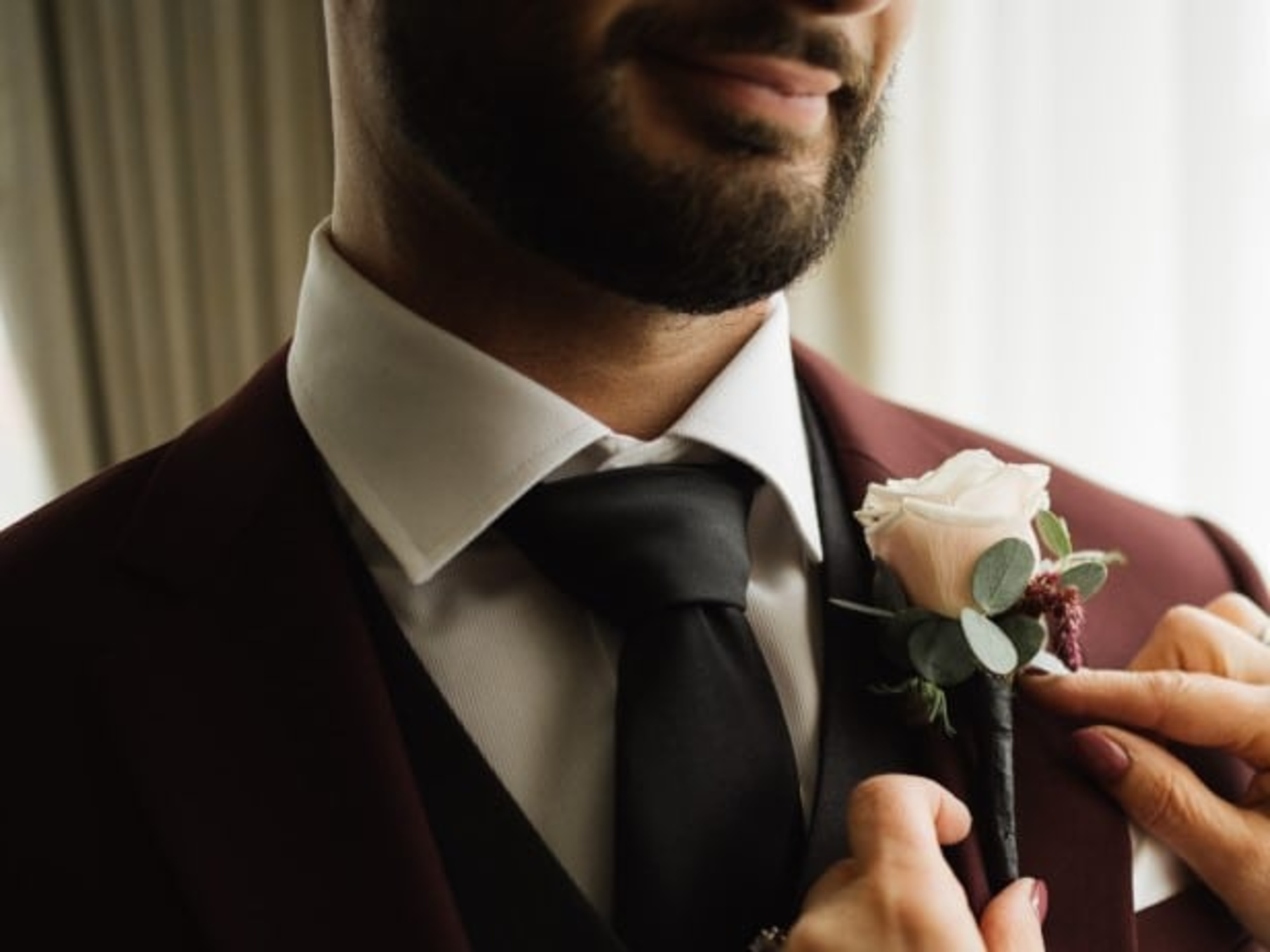 Close-up of a groom in a maroon suit as someone pins a blush rose boutonniere to his lapel.