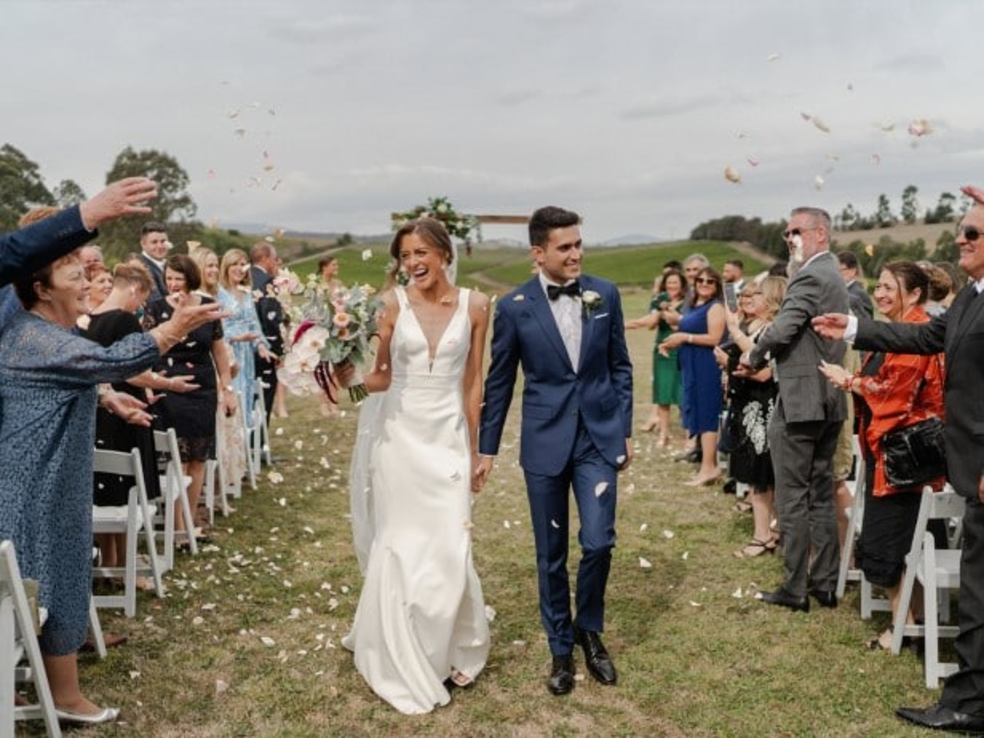 Bride and groom walk down an outdoor aisle smiling as guests toss flower petals around them.