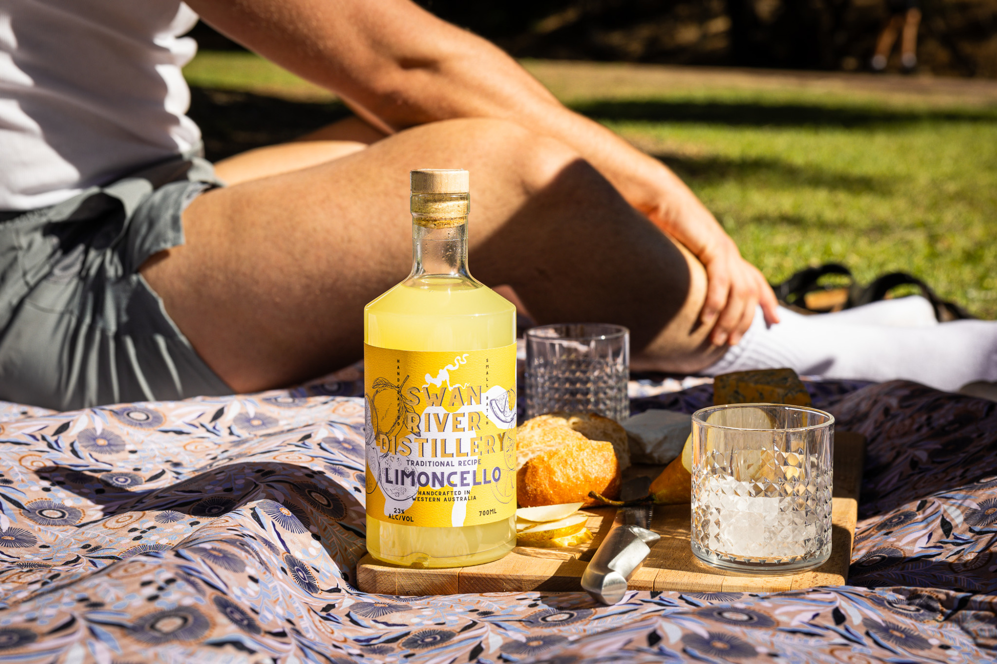 Bottle of limoncello with glasses and bread on a patterned picnic blanket in the sun.