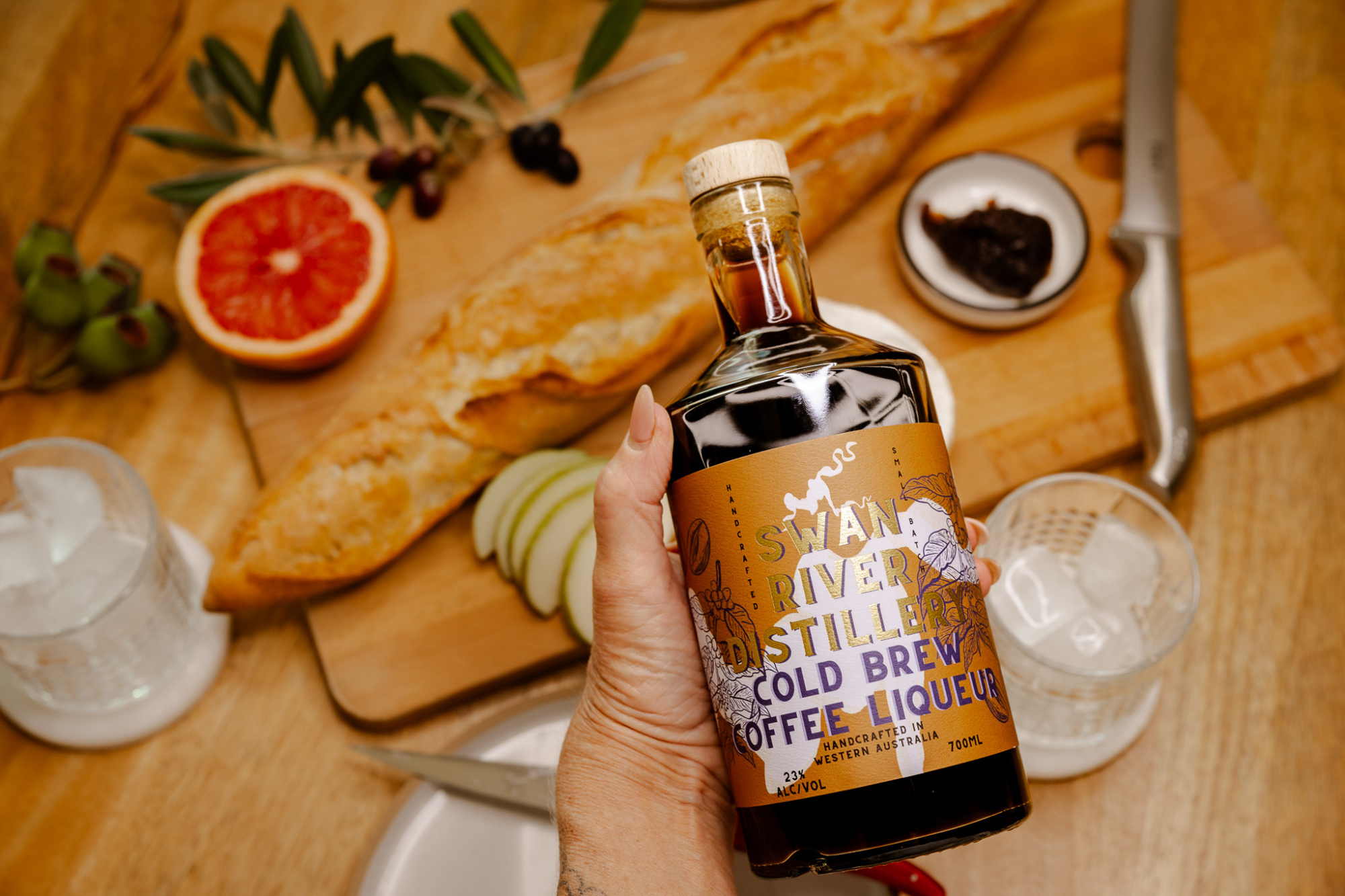 Hand holding a bottle of cold brew coffee liqueur above a rustic spread of bread, fruit, and barware.