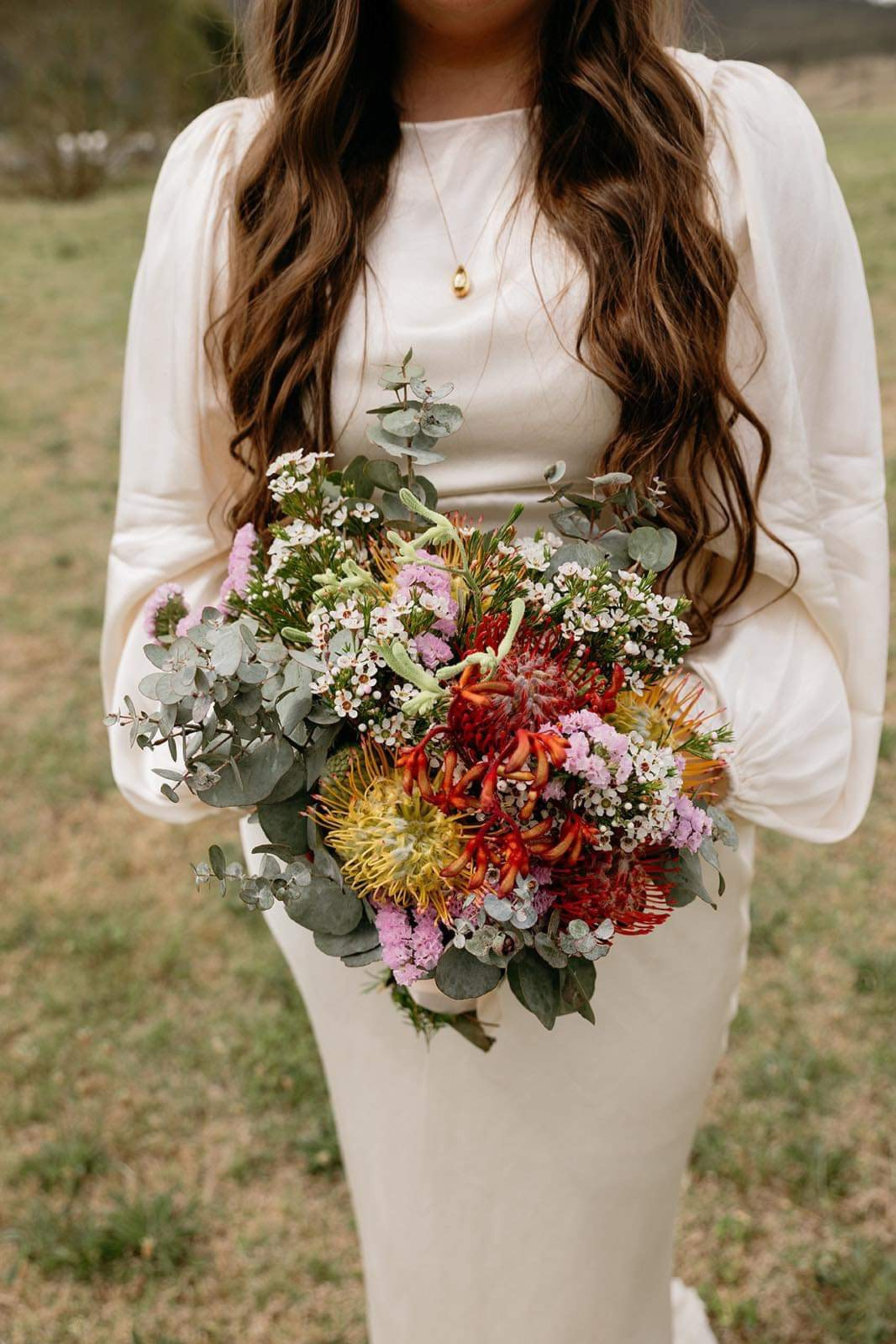 Bride in a long-sleeve ivory dress holding a colorful wildflower bouquet with greenery outdoors.