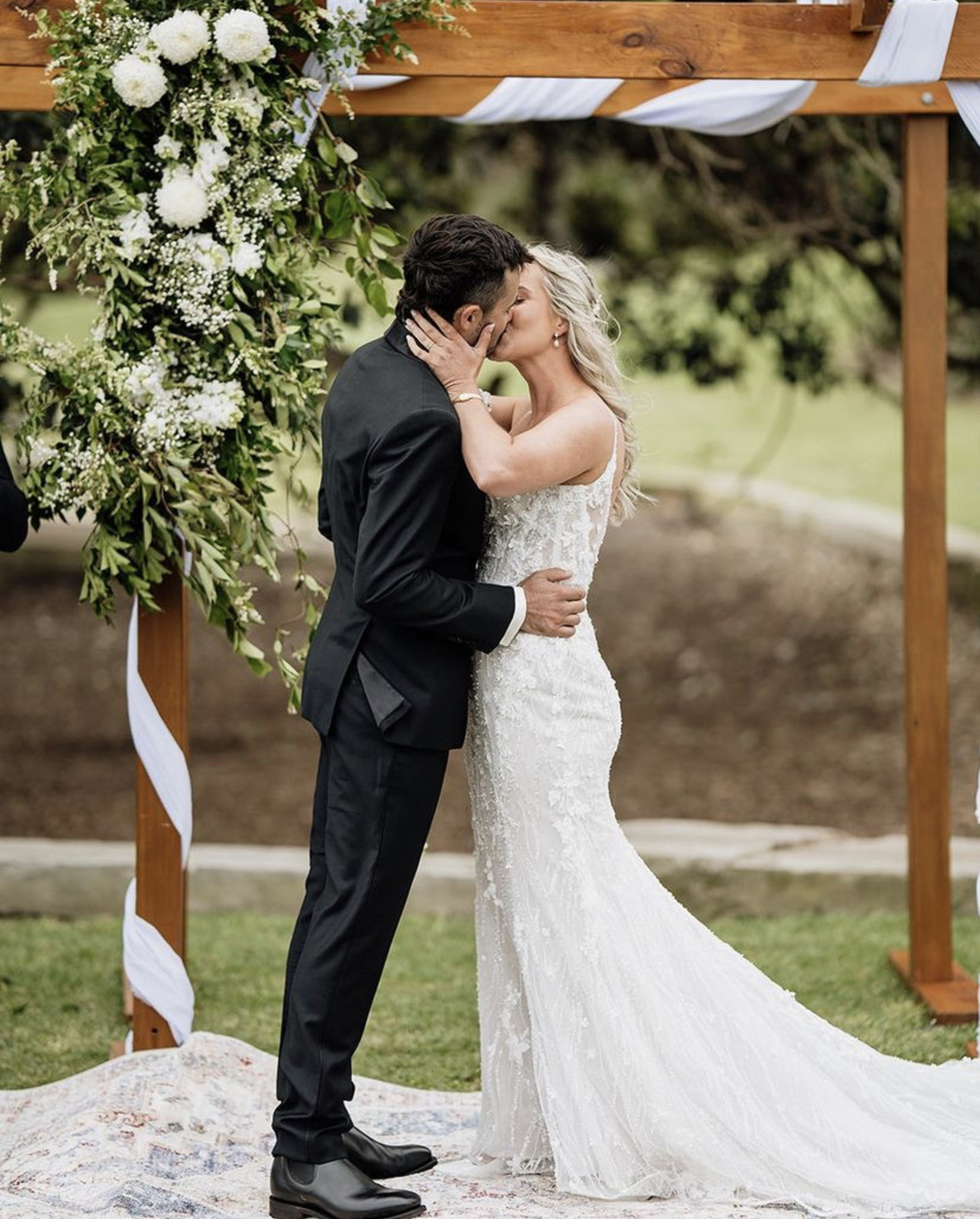 Bride and groom share a kiss under a floral wedding arch during an outdoor ceremony.