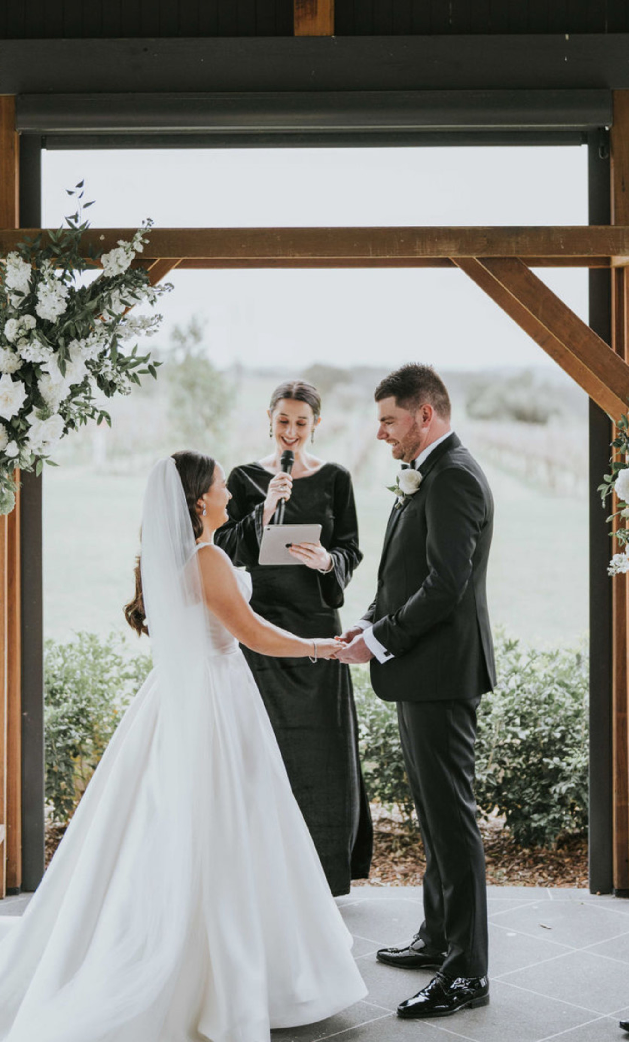 Couple holding hands during an outdoor wedding ceremony under a wooden arch with white floral arrangements.