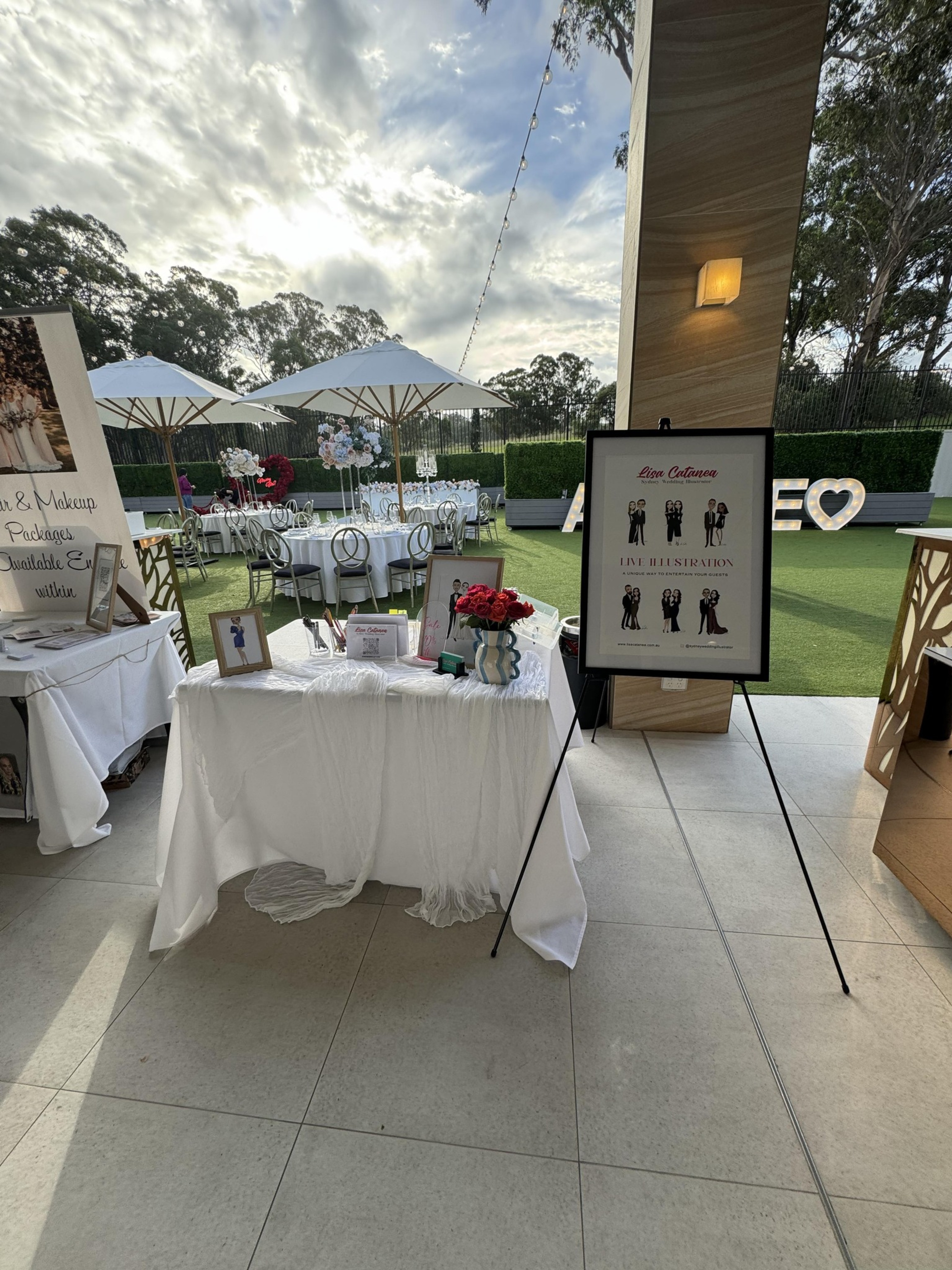 Outdoor wedding vendor display table with live illustration sign and decorated reception tables under white umbrellas.