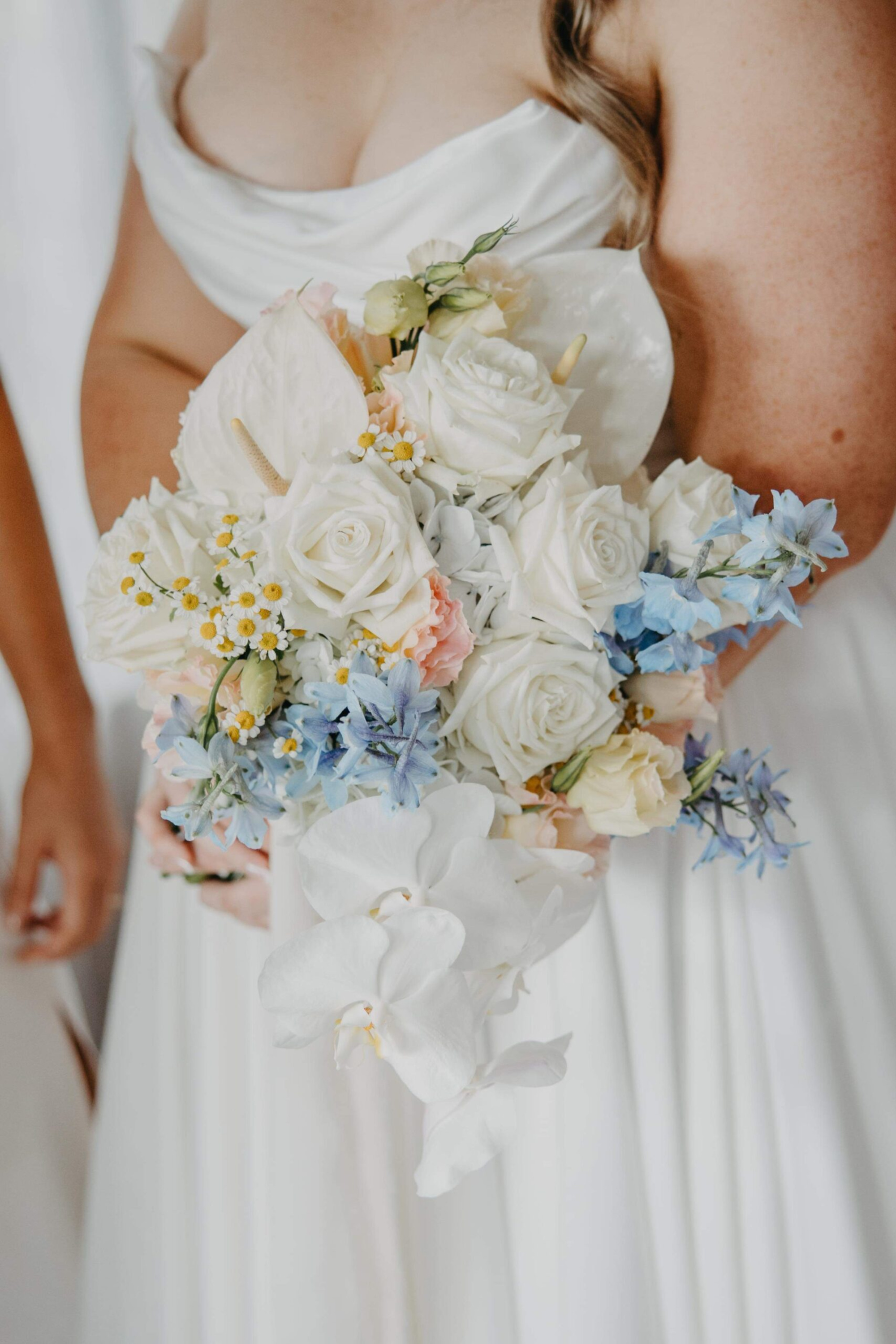 Bride holding a romantic pastel bouquet of white roses, orchids, and blue flowers against her white wedding dress.