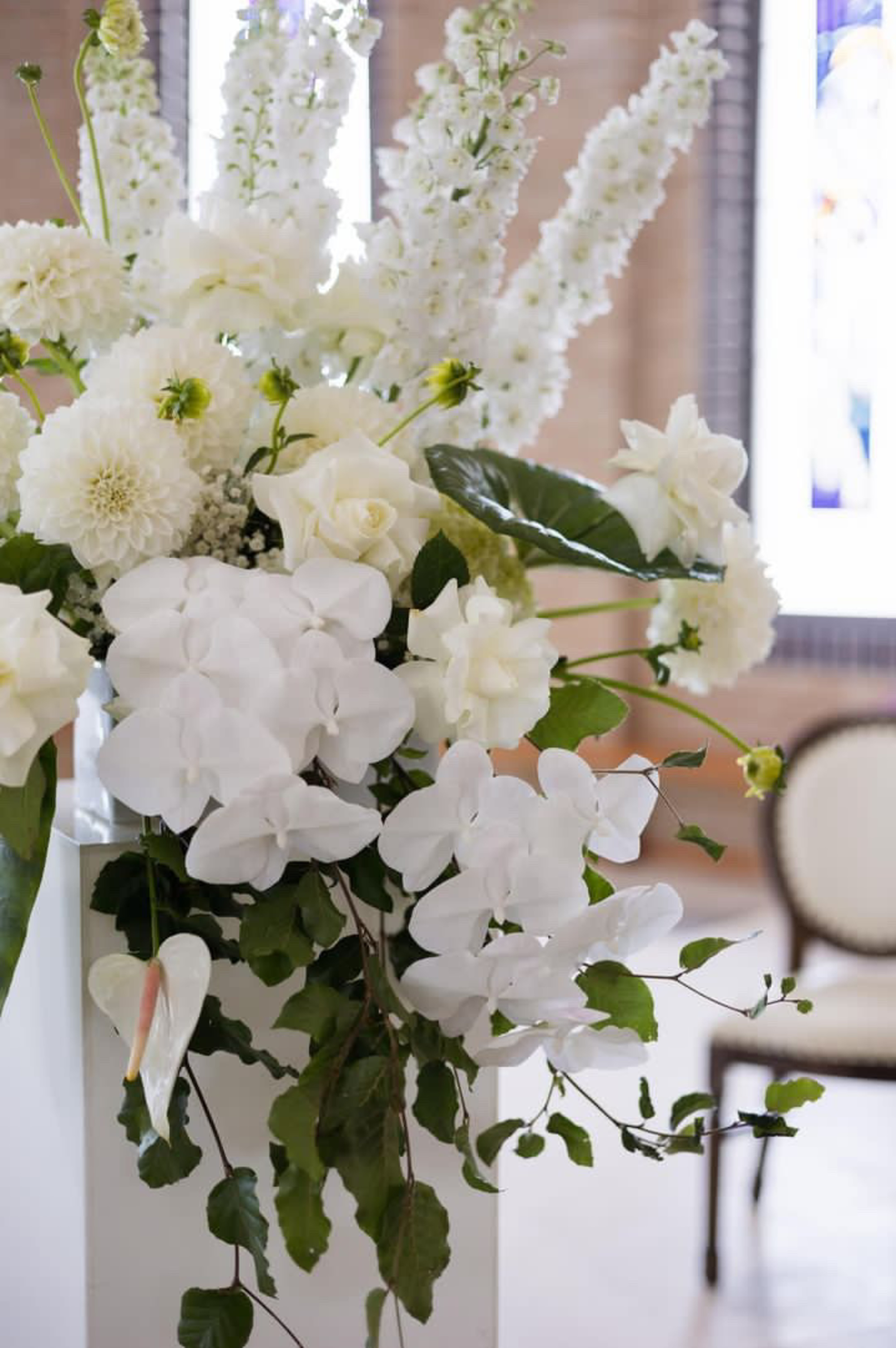 Elegant white wedding flower arrangement with orchids, roses, and greenery on a pedestal indoors.