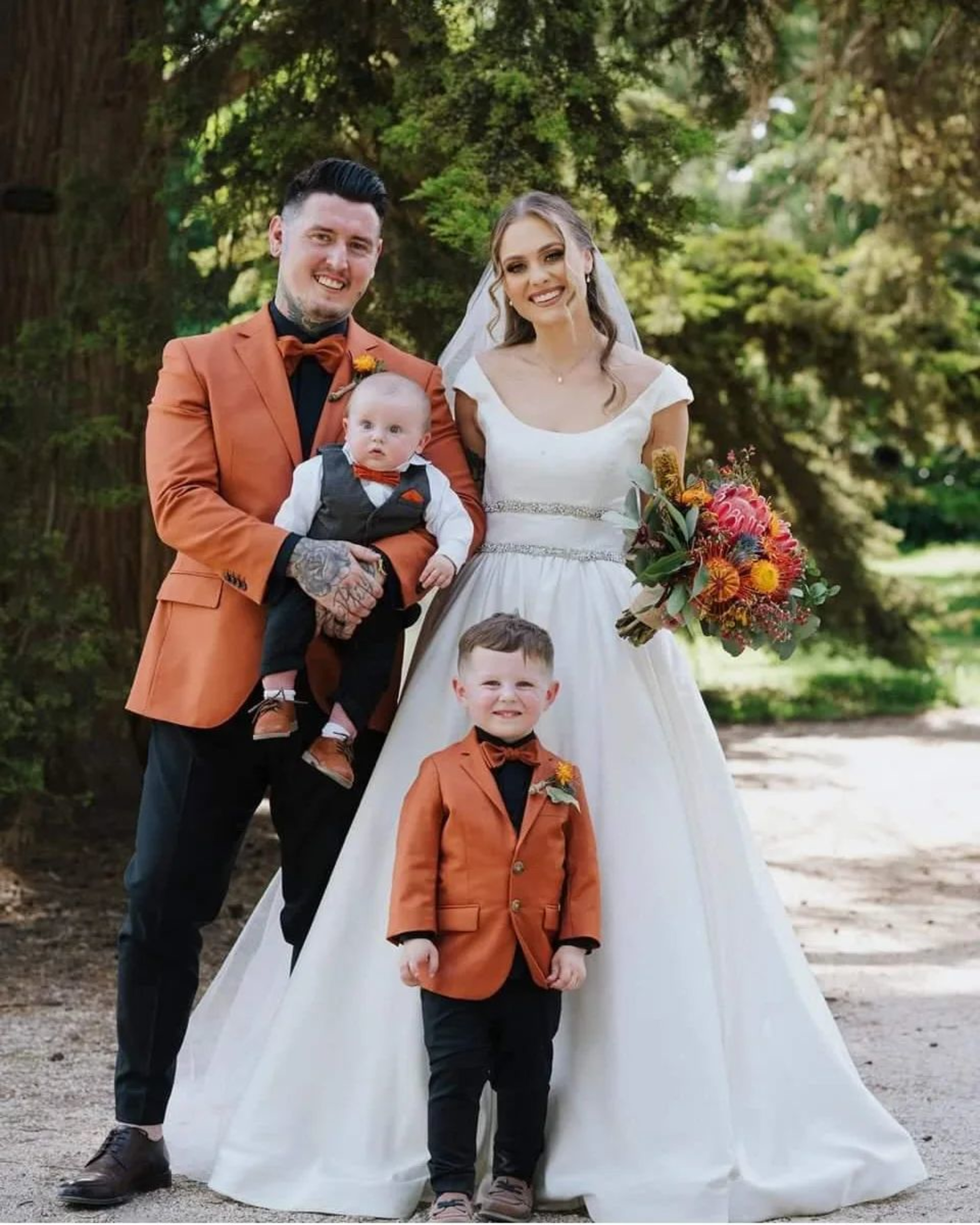 Outdoor wedding family portrait with bride in white gown and groom and sons in matching orange jackets.