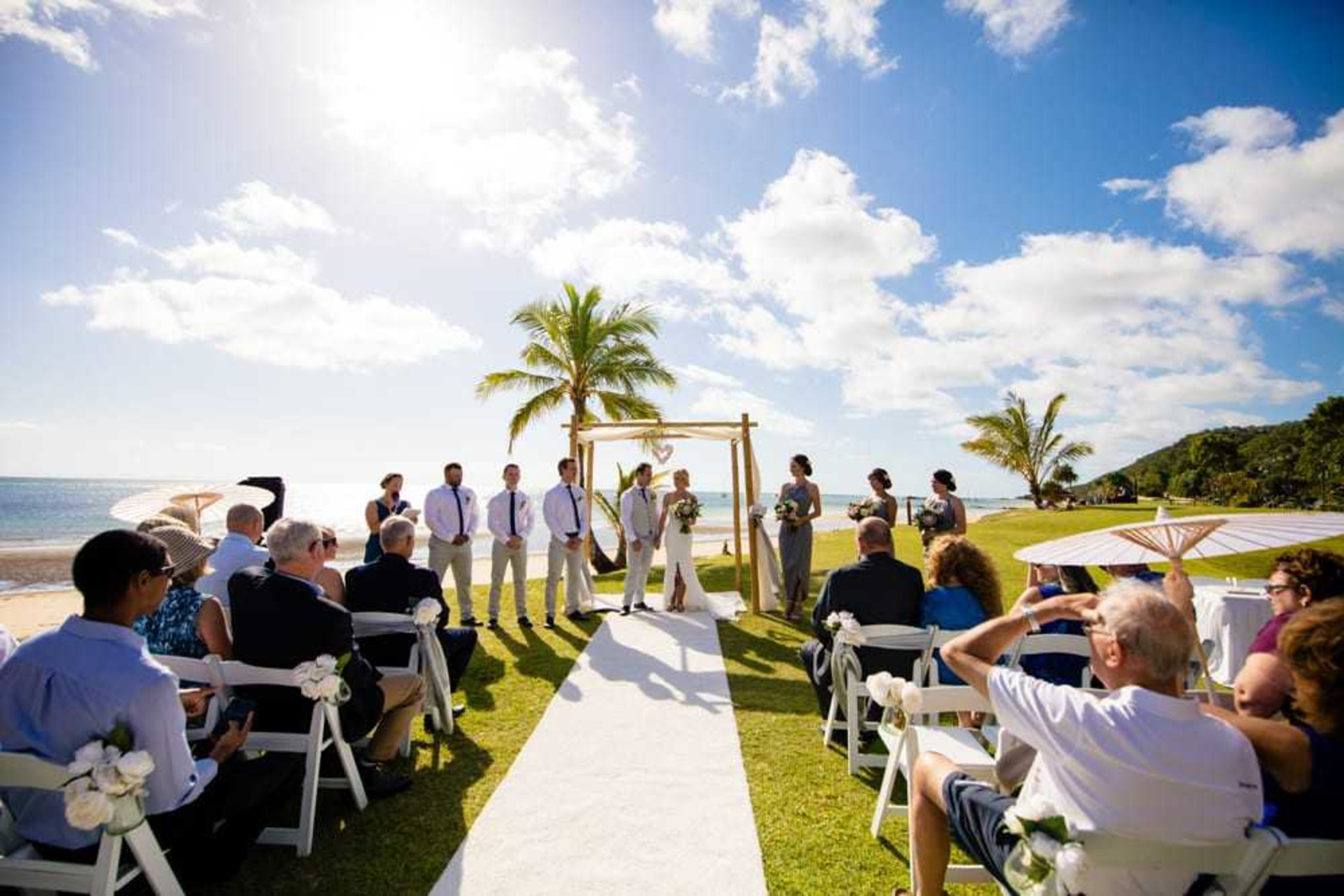 Outdoor beach wedding ceremony with guests seated along an aisle facing a couple under a wooden arch by the ocean.