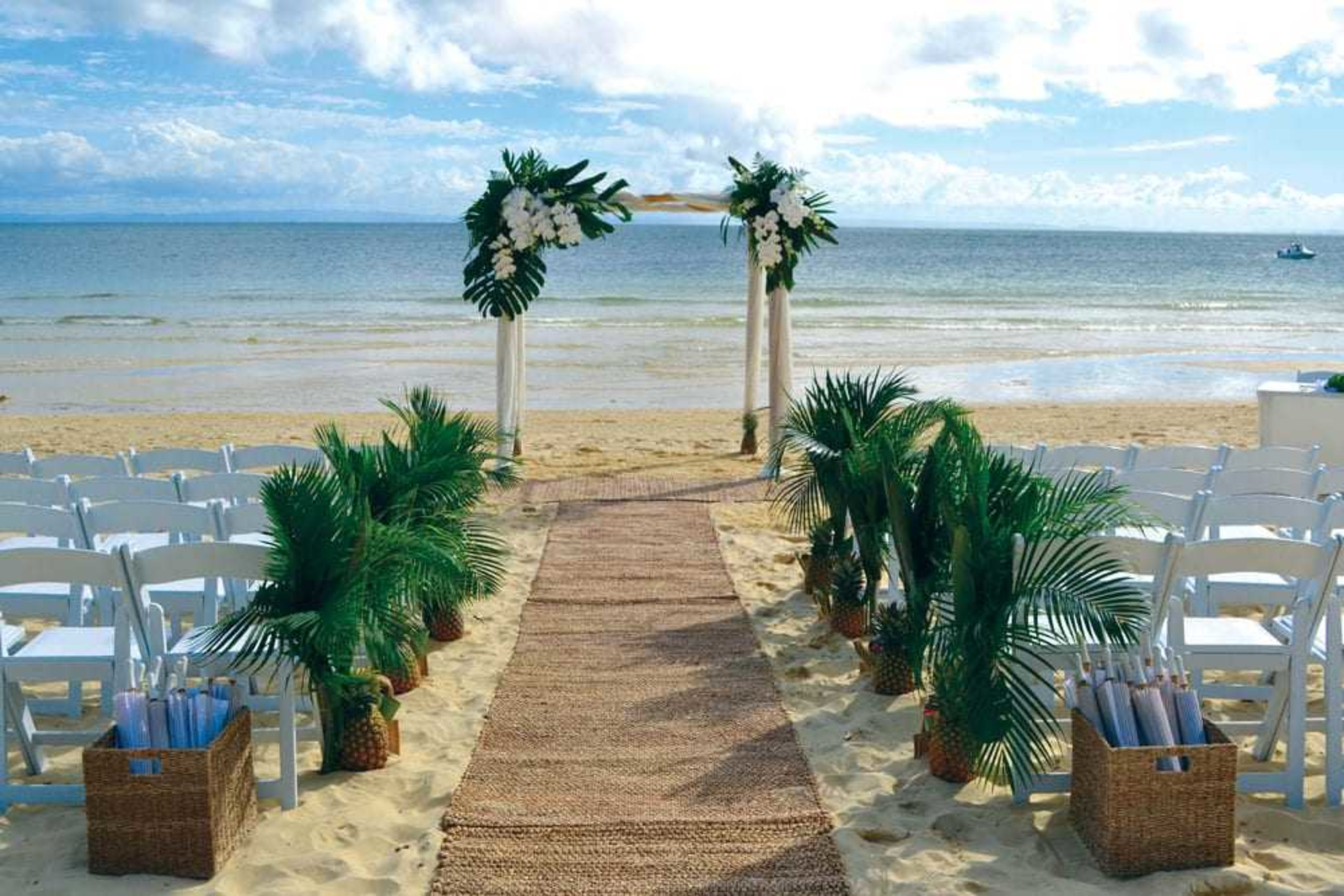 Tropical beach wedding ceremony setup with white chairs, palm decor, and a floral arch facing the ocean.