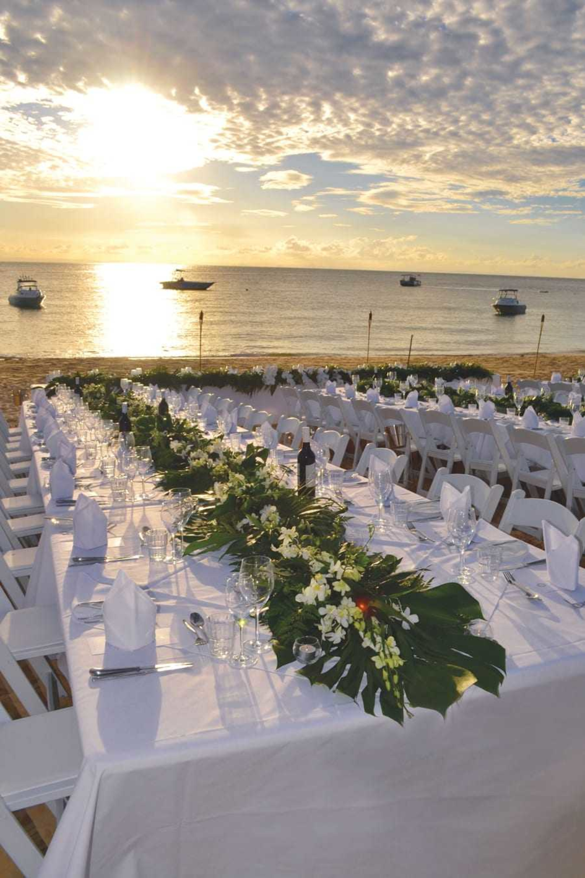 Elegant wedding reception tables set on a beach at sunset with tropical greenery centerpieces overlooking the ocean.