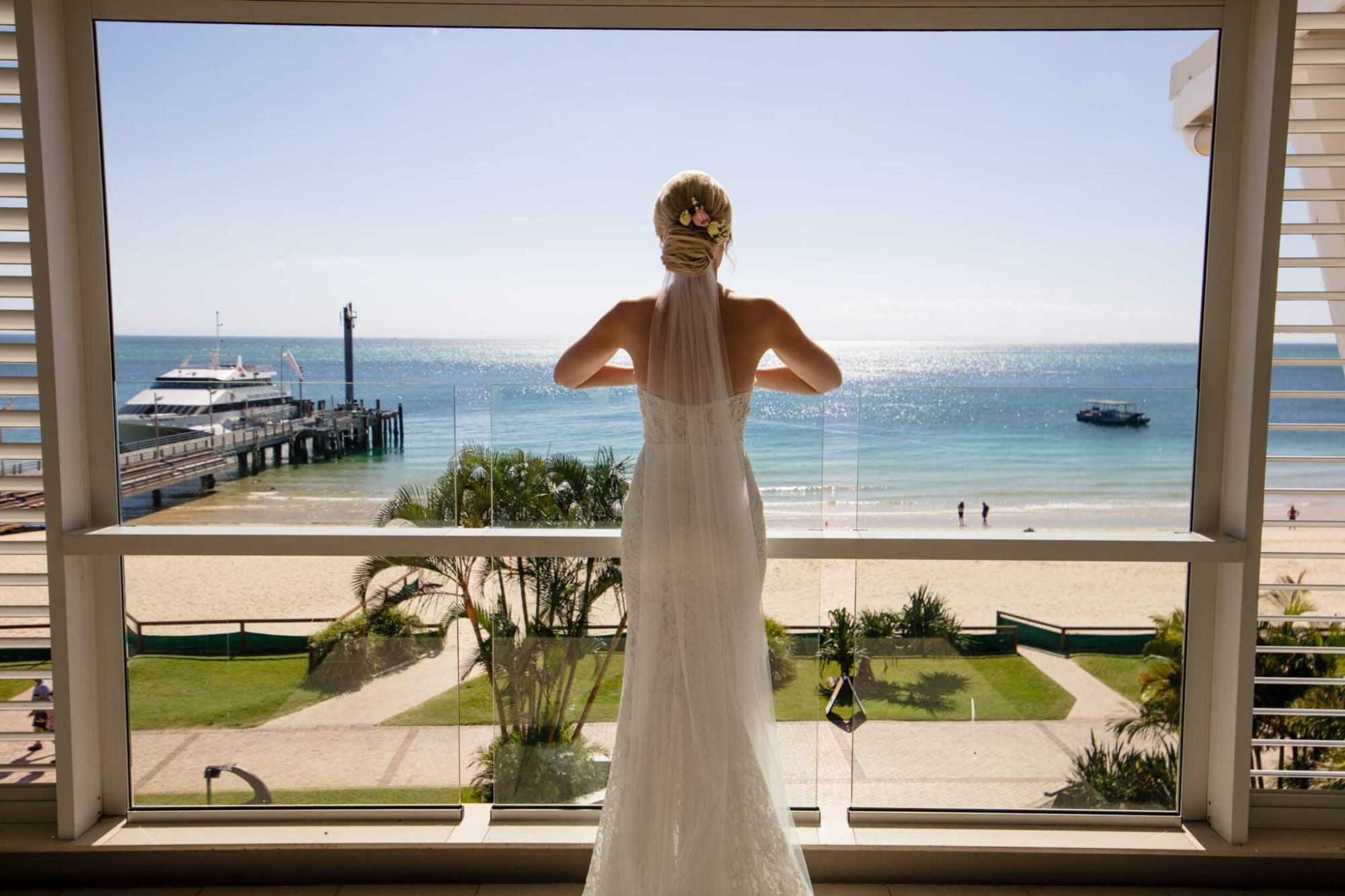 Bride in a lace gown looks out over a sunny beach and ocean from a waterfront venue balcony.