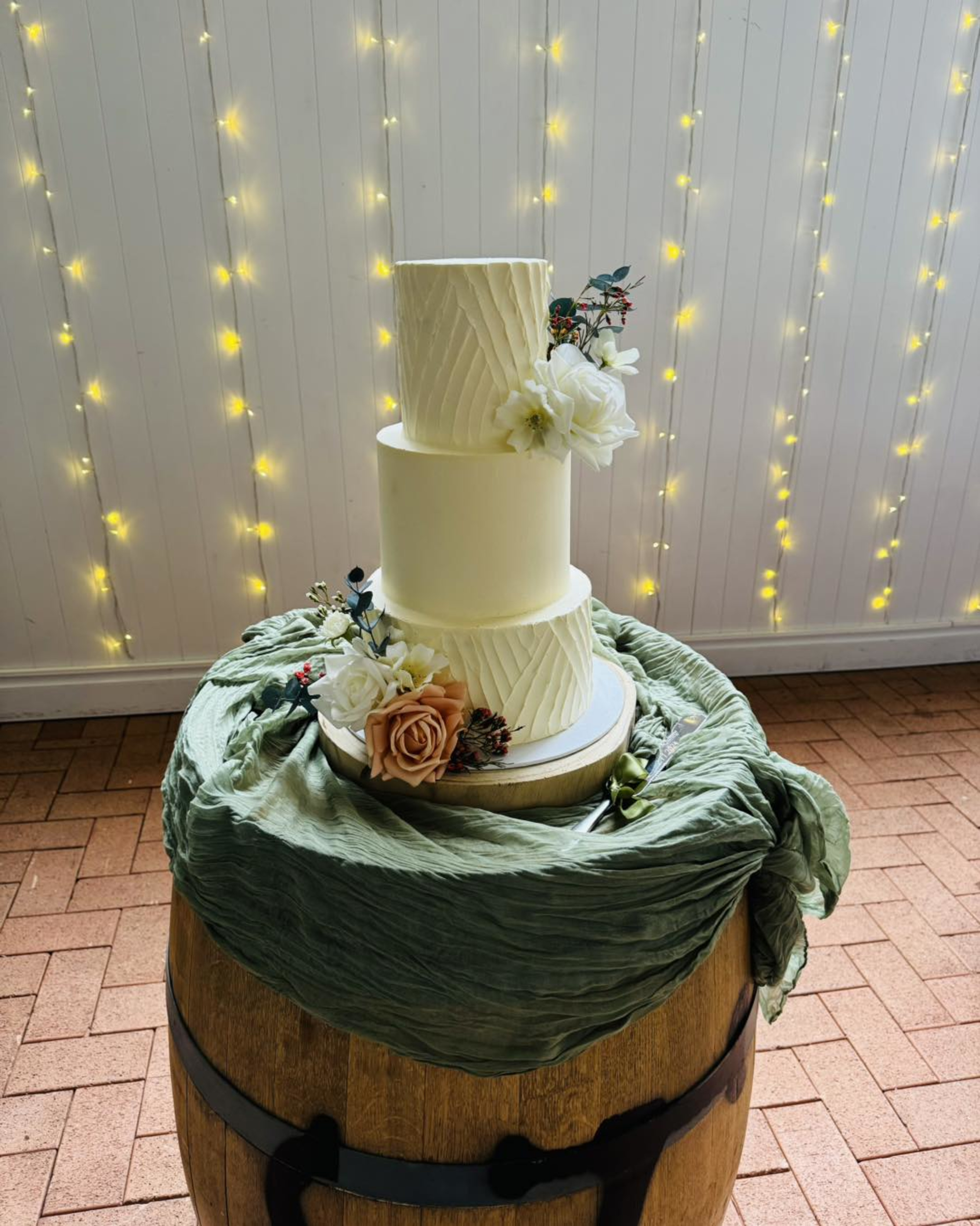 Three-tier white wedding cake with floral accents on a rustic barrel stand draped in green fabric and fairy lights behind.
