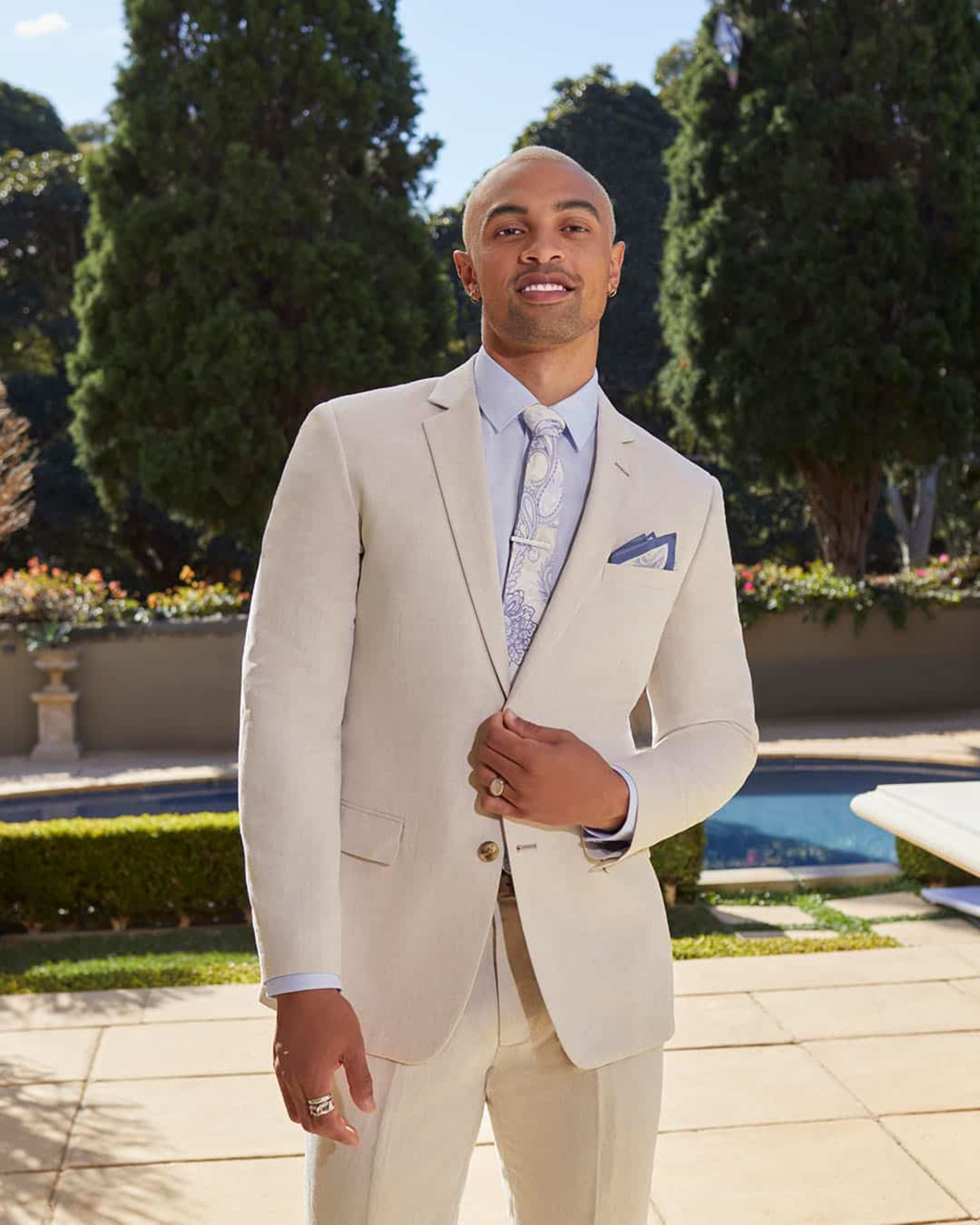 Groom wearing a light beige suit with patterned tie posing in an elegant garden venue.