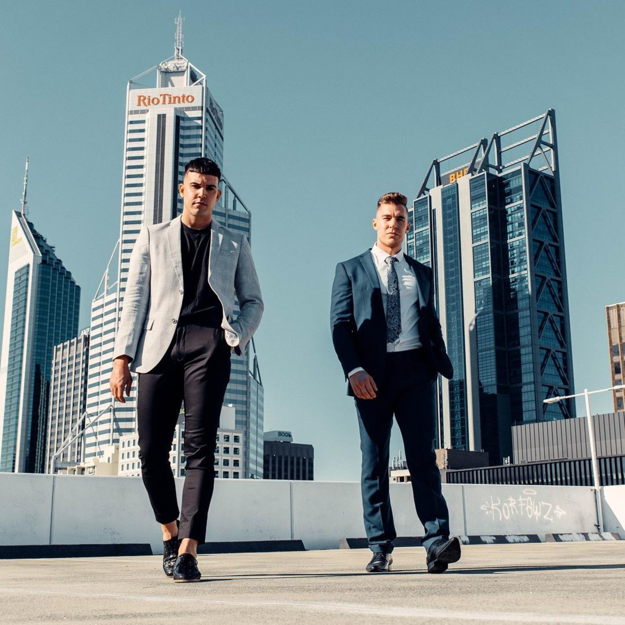 Two men in stylish suits walk across a rooftop with modern city skyscrapers in the background.