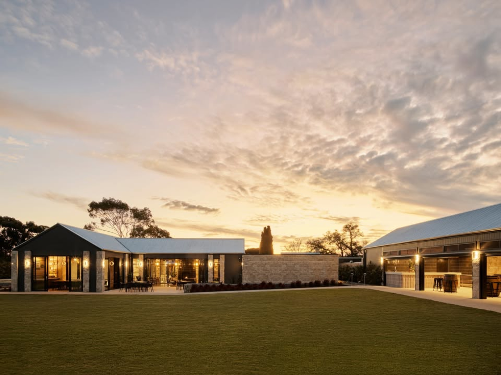 Modern barn-style wedding venue at sunset with large lawn and warm glowing lights.