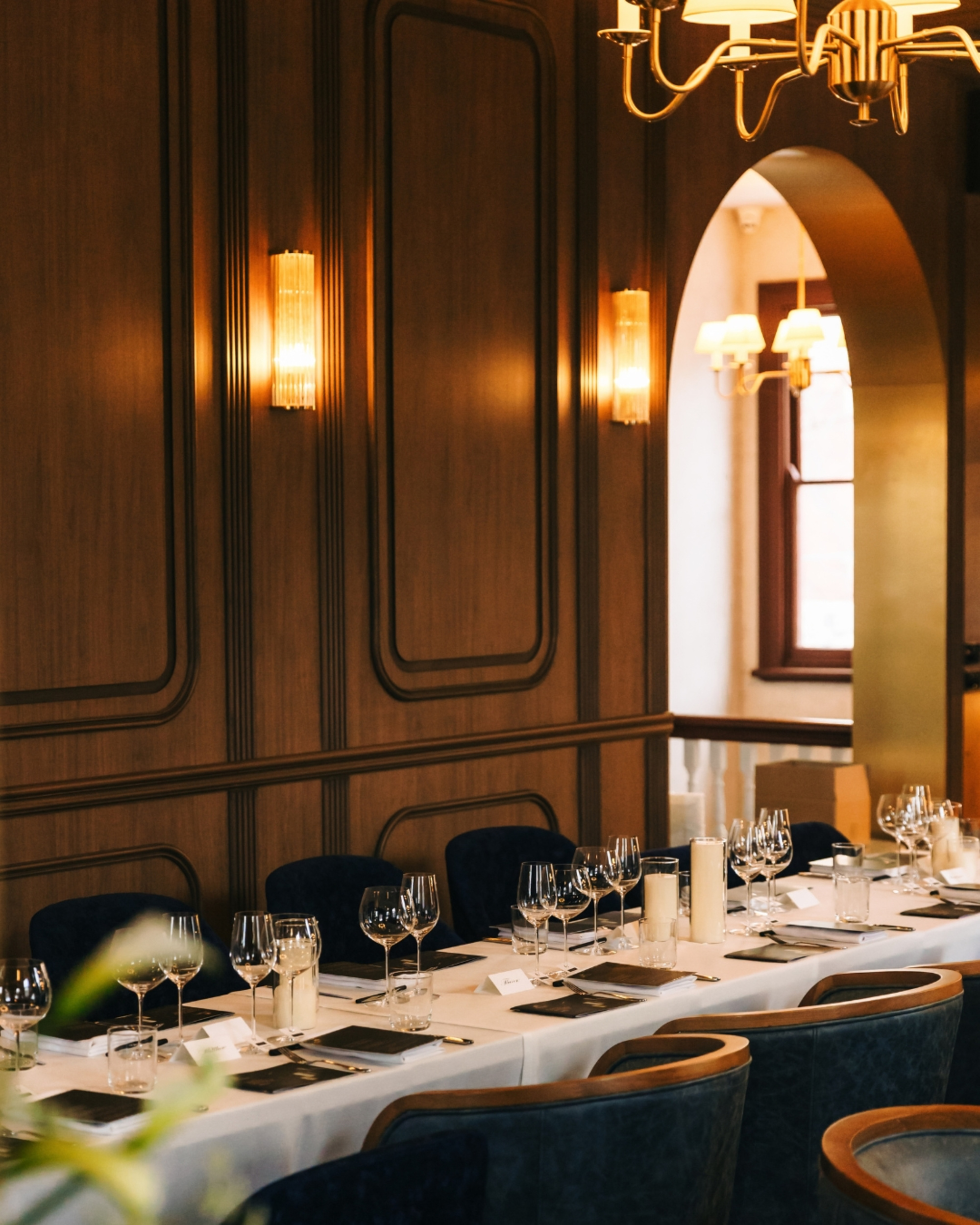 Elegant indoor wedding reception table set with glassware, candles, and place cards in a warmly lit wood-paneled room.