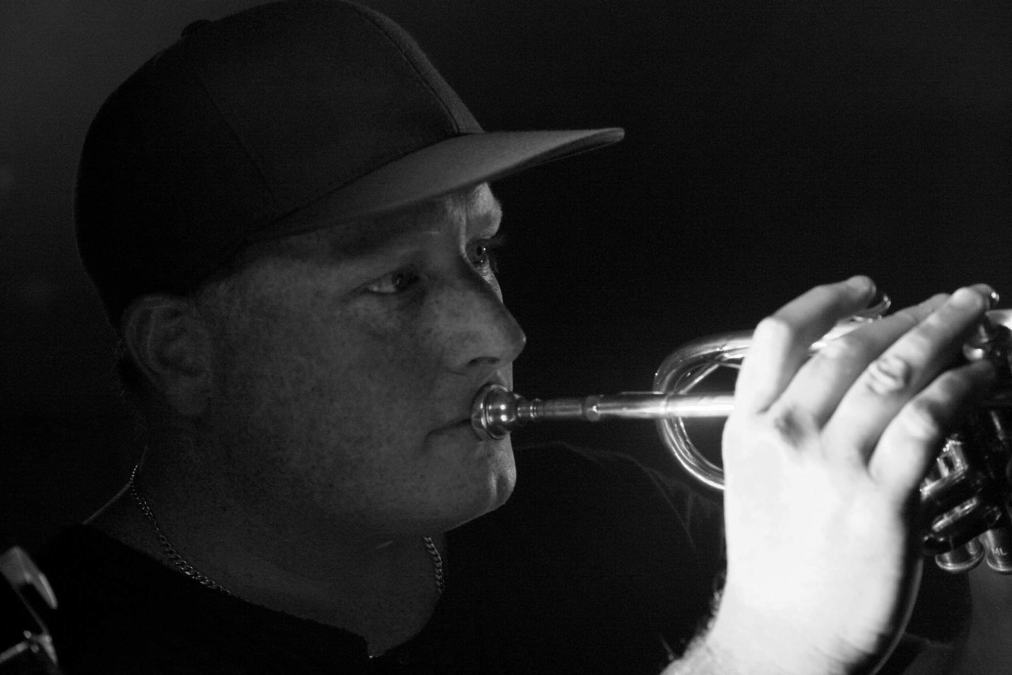Black and white close-up of a trumpet player performing, lit dramatically against a dark background.