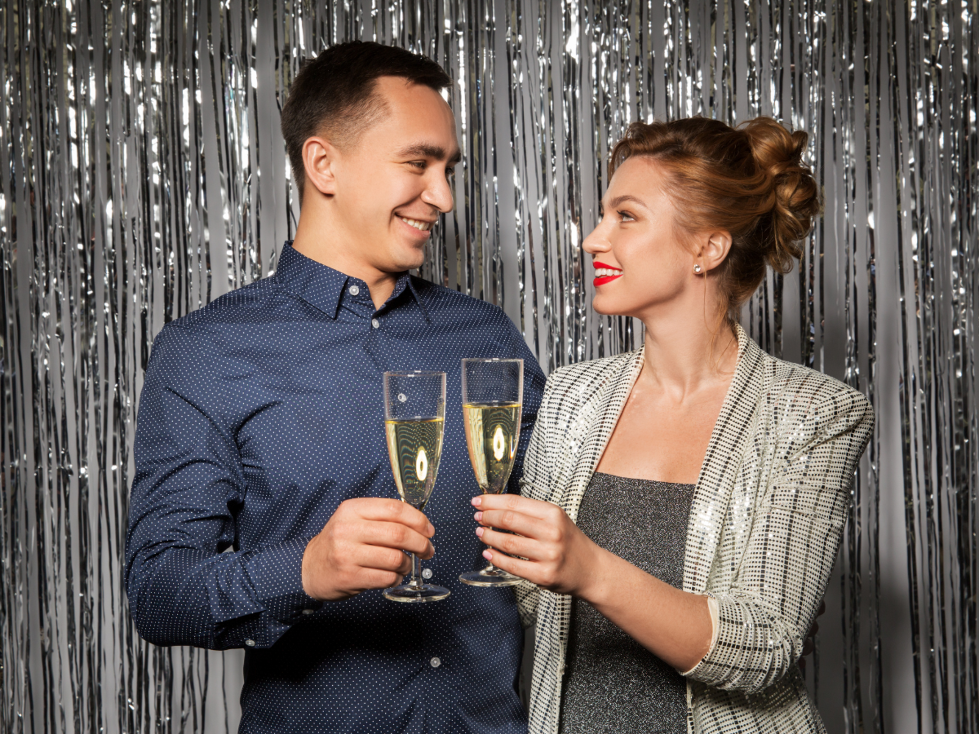 Smiling couple clinks champagne glasses in front of a silver tinsel backdrop.