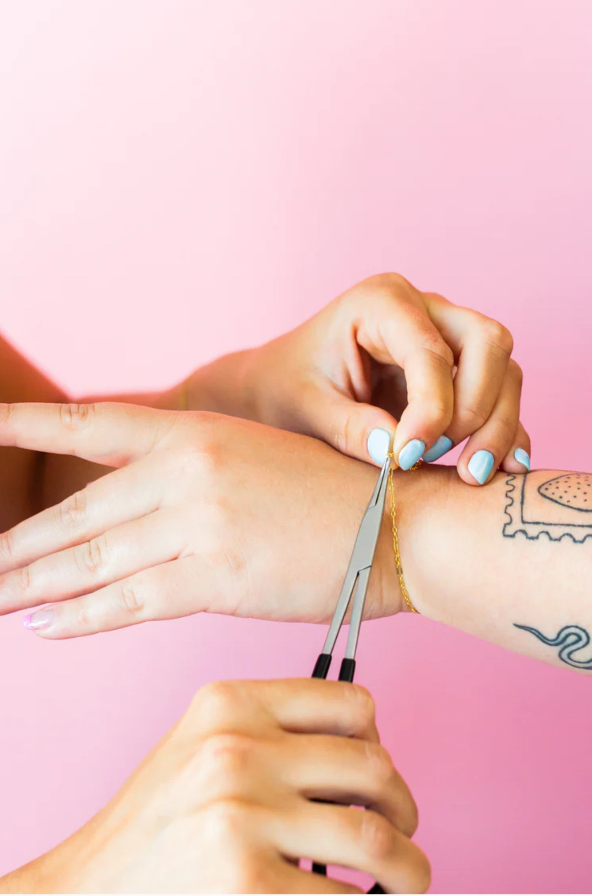 Hands securing a delicate gold bracelet on a wrist against a pink background