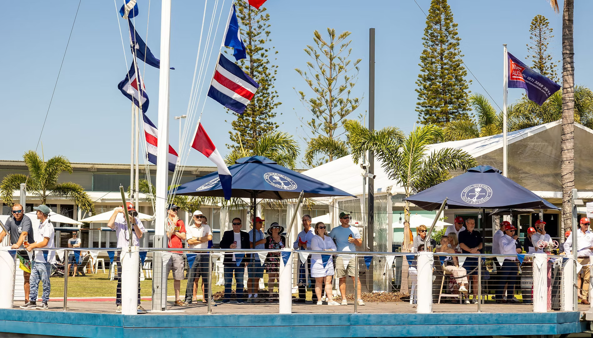 Guests gather on a sunny waterfront deck with nautical flags and umbrellas at a marina-style venue.