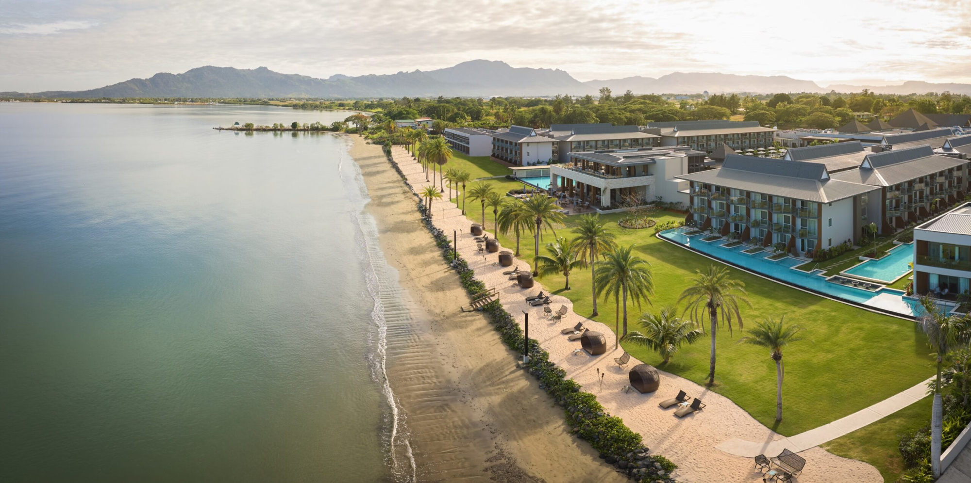 Aerial view of a luxury beachfront resort with palm trees, pools, and a sandy shoreline beside calm ocean water.