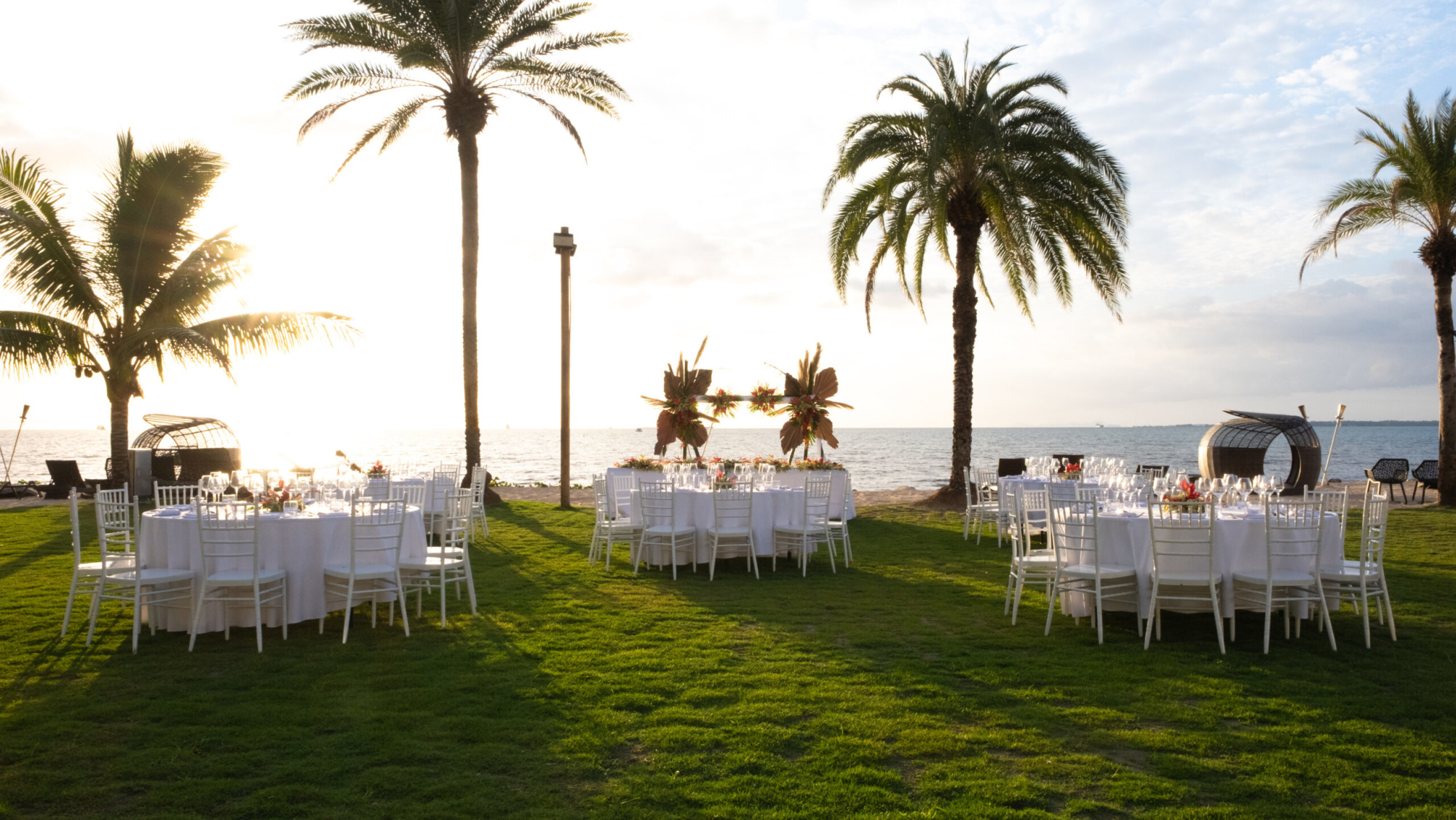 Sunset beach wedding reception setup with round tables and white chairs under palm trees by the ocean.