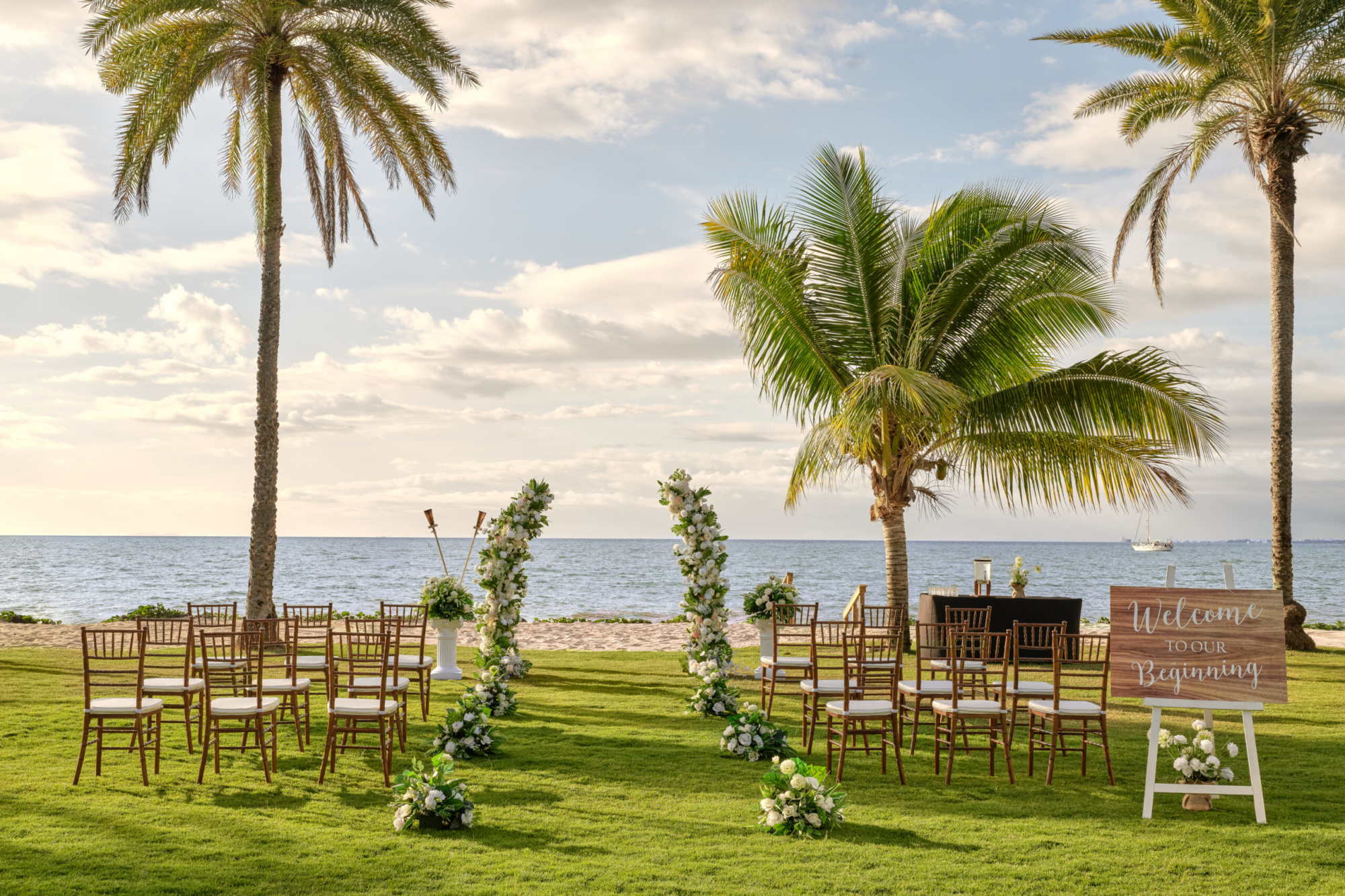 Oceanfront tropical wedding ceremony setup with floral arches, wooden chairs, and palm trees at sunset.