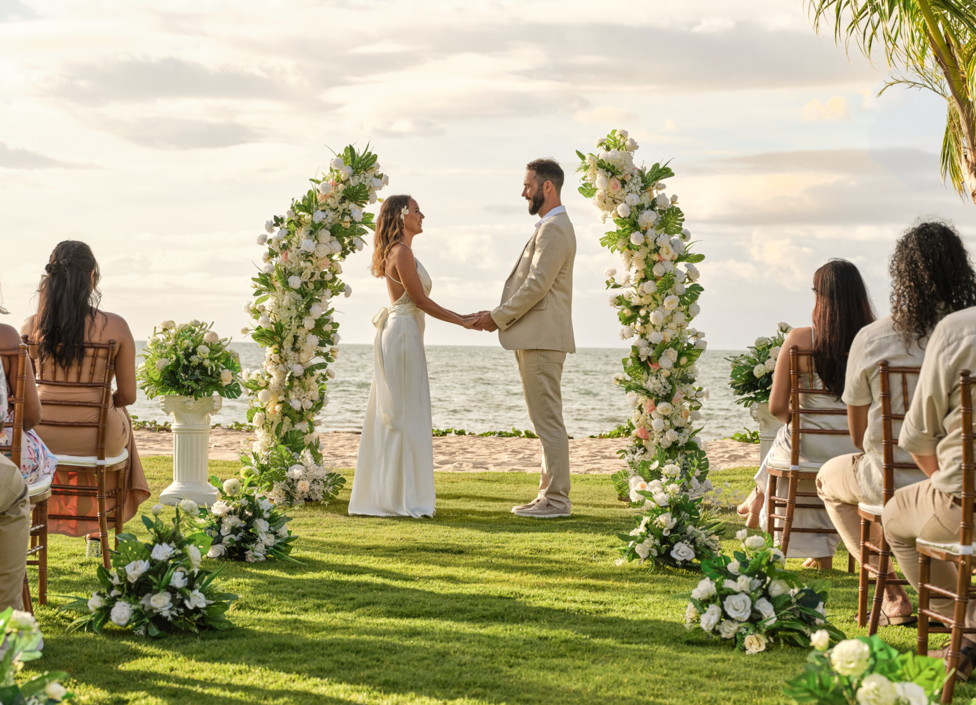 Couple holding hands under a floral arch during an intimate beach wedding ceremony by the ocean.